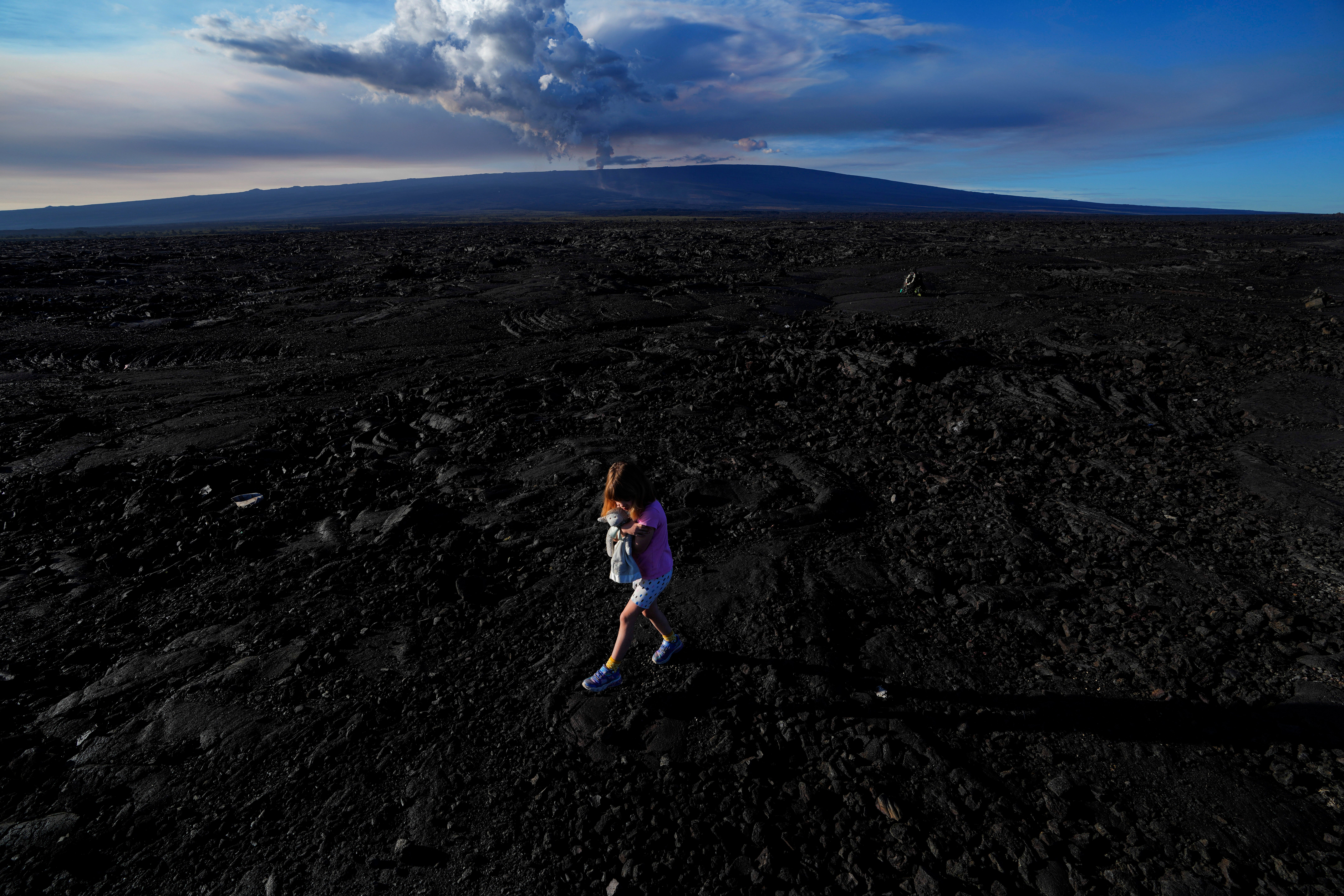 Abigail Dewar, of Alberta, Canada, holds a stuffed animal as she walks over hardened lava rock from a previous eruption as the Mauna Loa volcano erupts, behind, Wednesday, Nov. 30, 2022, near Hilo, Hawaii