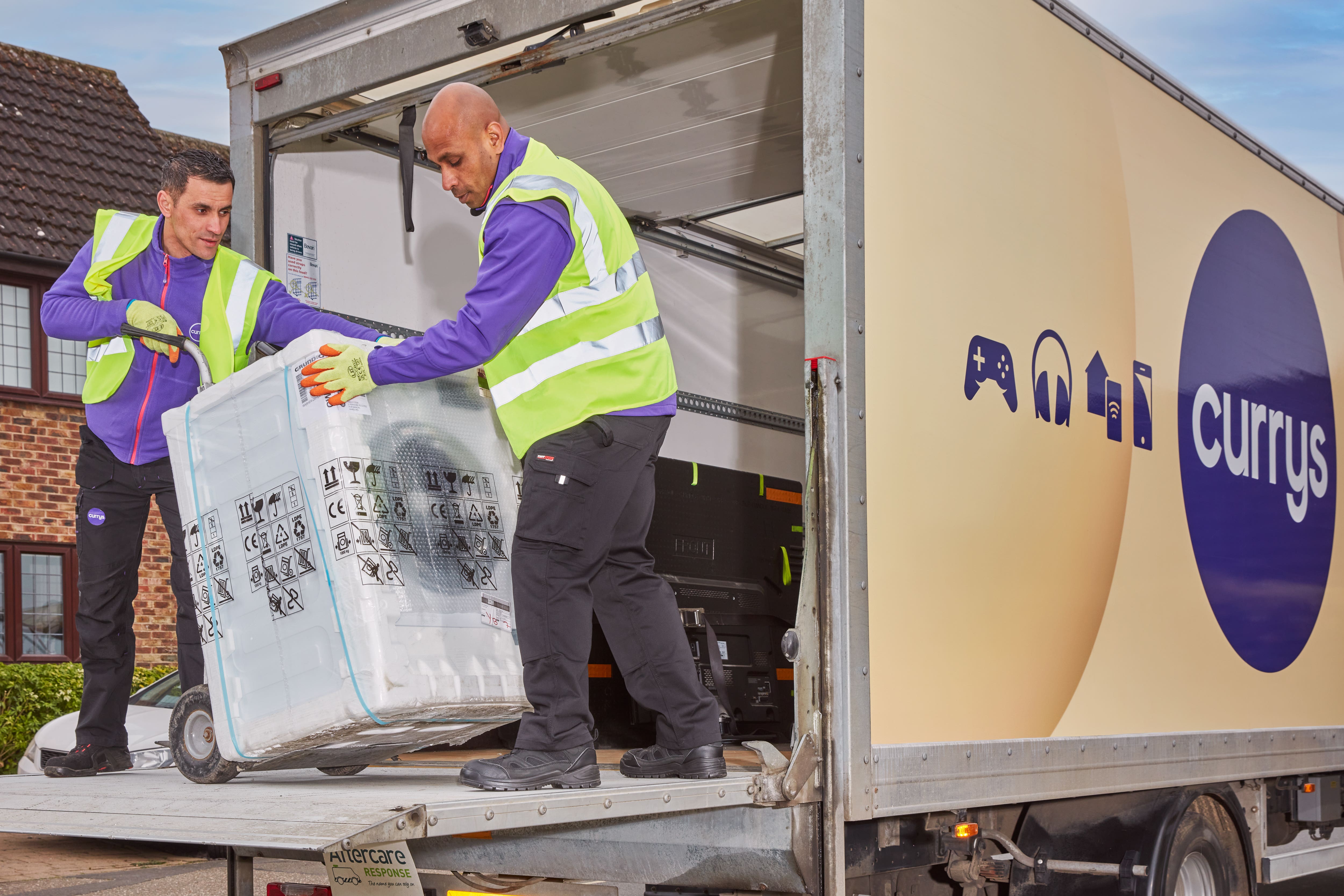  Two men in purple work uniforms unload a large box labelled 'aftercare response' from the back of a Currys delivery truck, with the text 'Currys' and images of gaming consoles and headphones on the side of the truck.