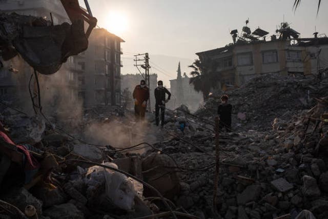 <p>Workers stand on top of a collapsed building as a digger works its way through the debris on 13 February 2023 in Hatay, Turkey</p>