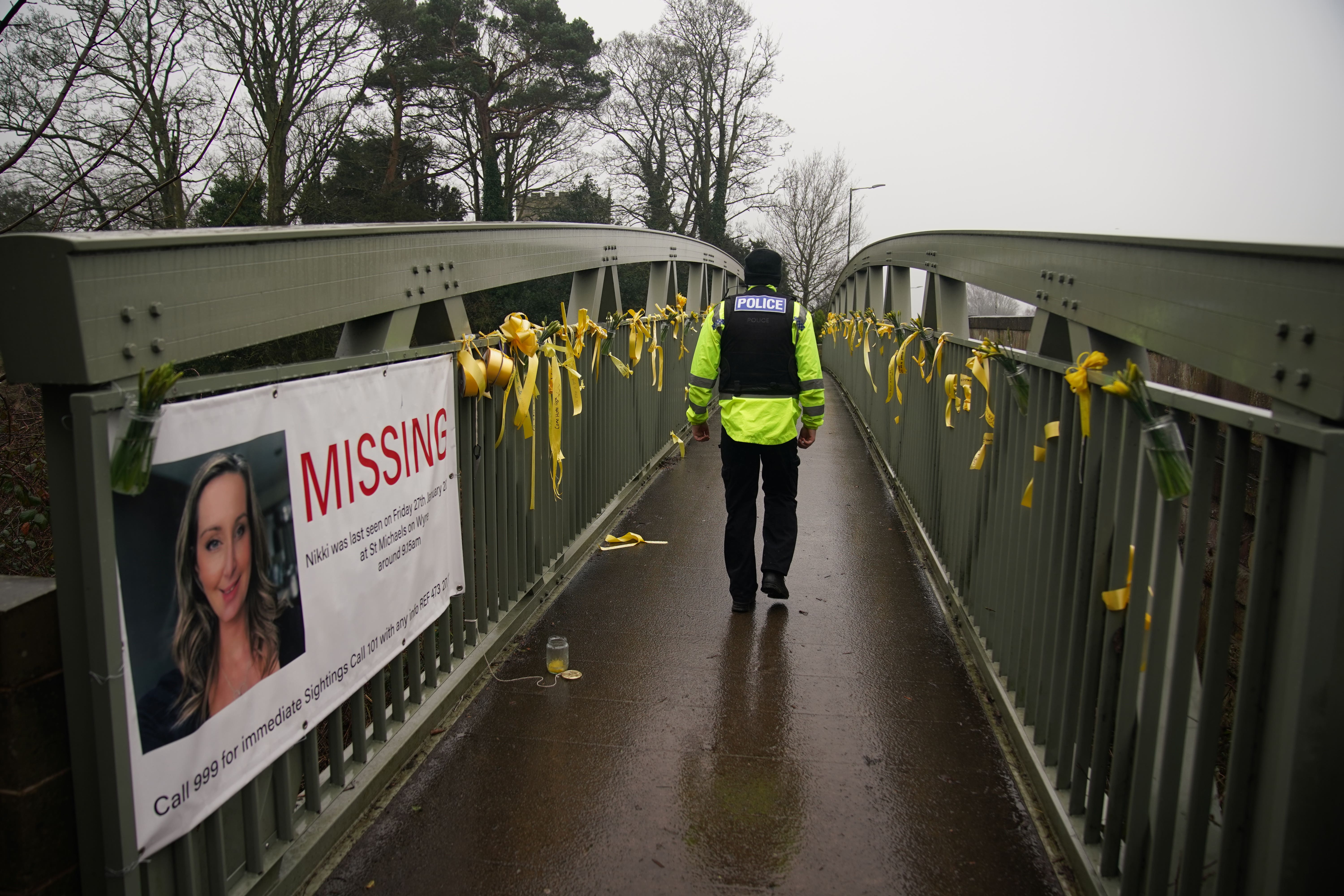 A police officer walks past a missing person appeal poster for Nicola Bulley and yellow ribbons and messages of hope tied to a bridge over the River Wyre