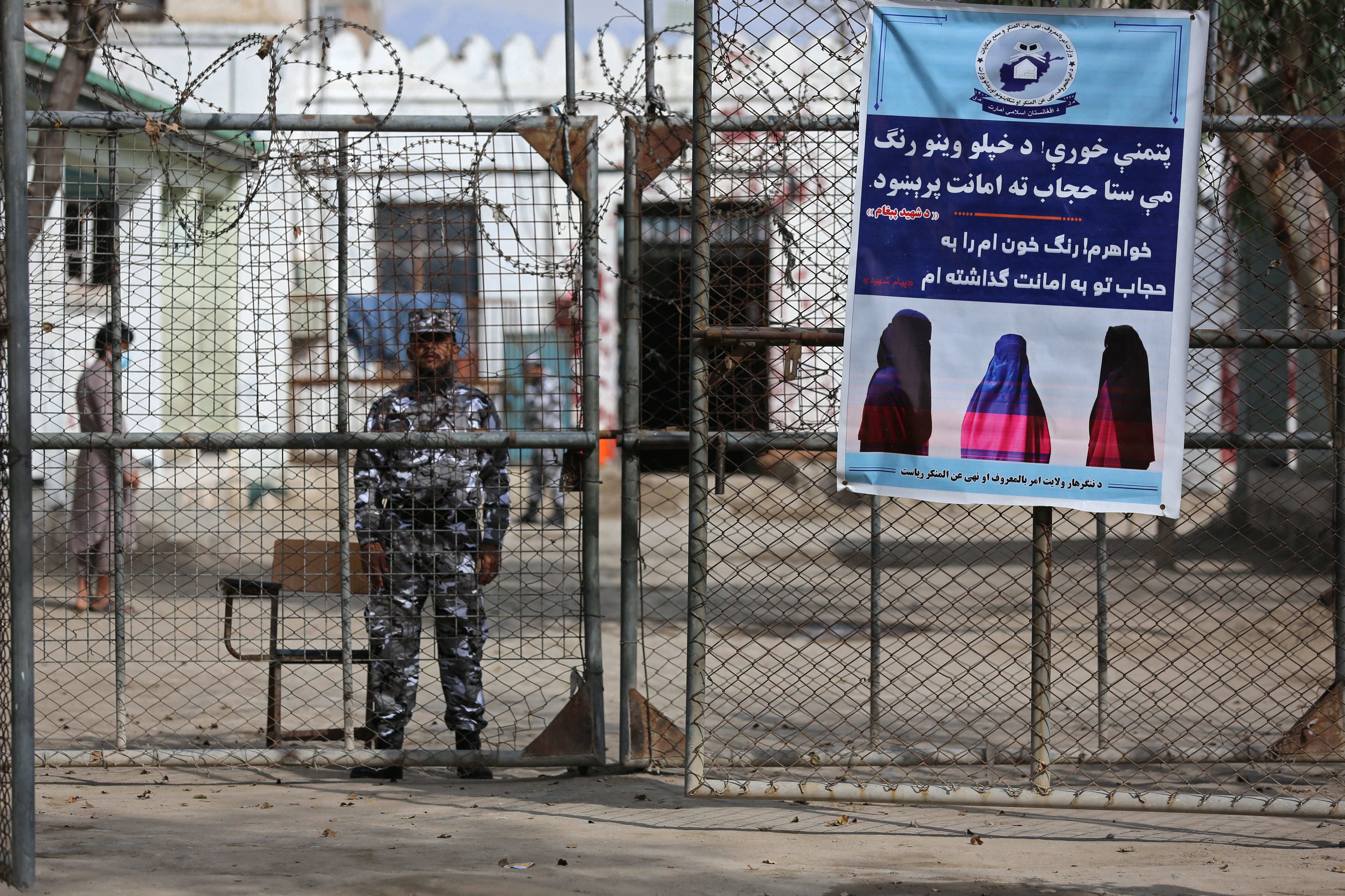 A Taliban security guard stands by a poster ordering women to wear Hijab, during a ceremony marking the distribution of new uniforms by the Taliban authorities at a prison in Jalalabad