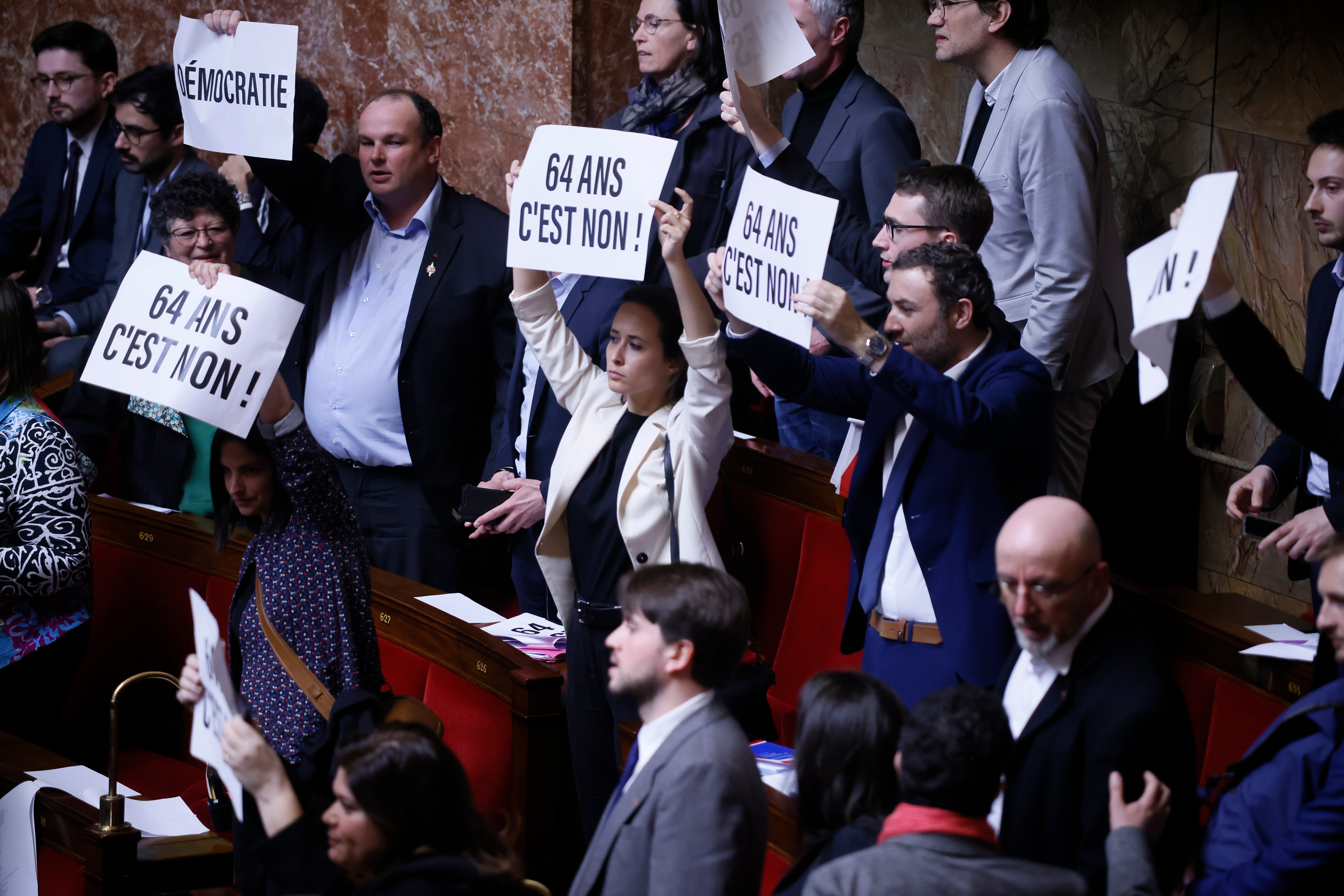 <p>Far-left legislators react as they hold papers reading: ‘64 years. It is no’ at the National Assembly in Paris</p>
