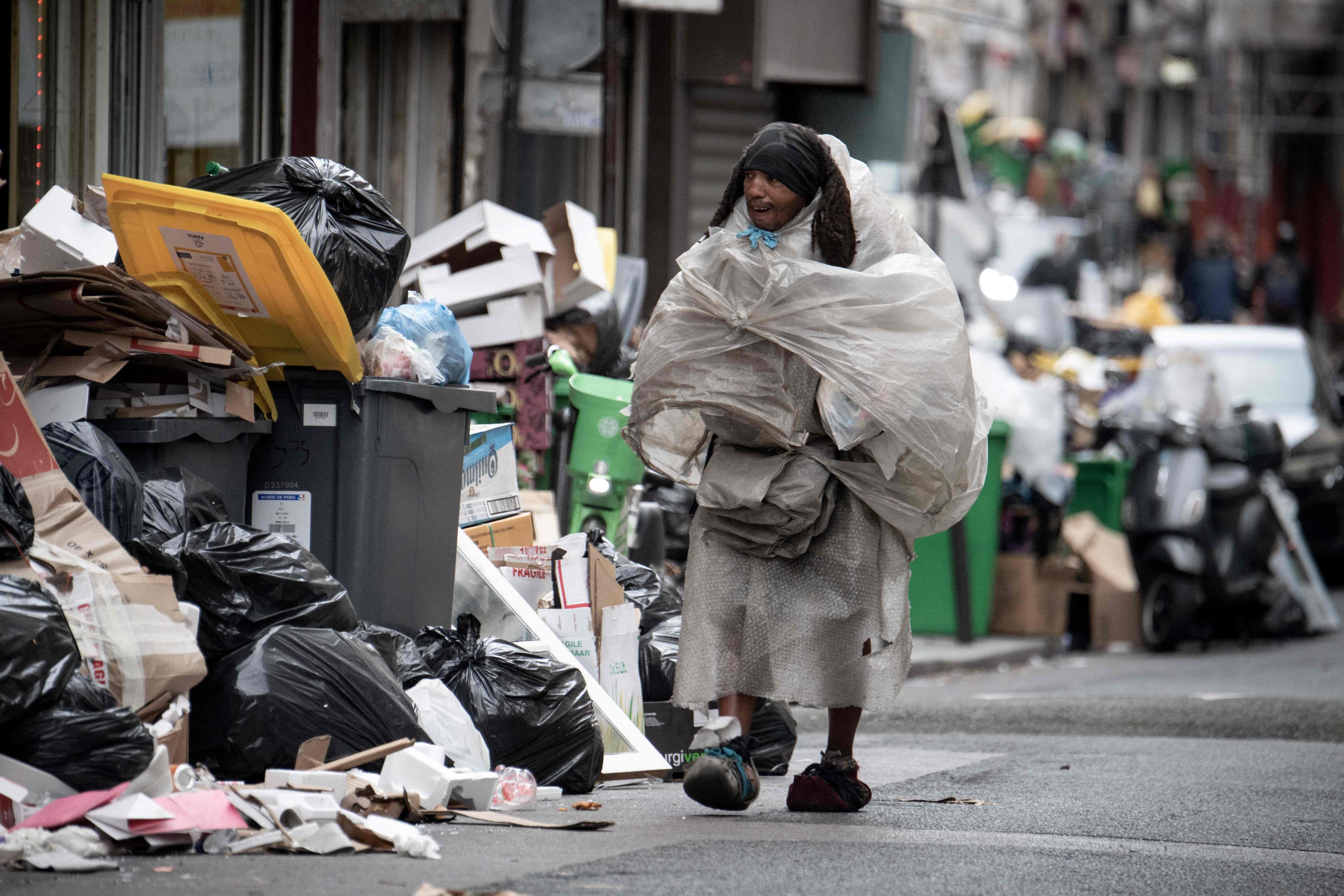 A homeless man, covered with plastic bags, walks past garbage bags that have been piling up on the pavement as waste collectors are on strike