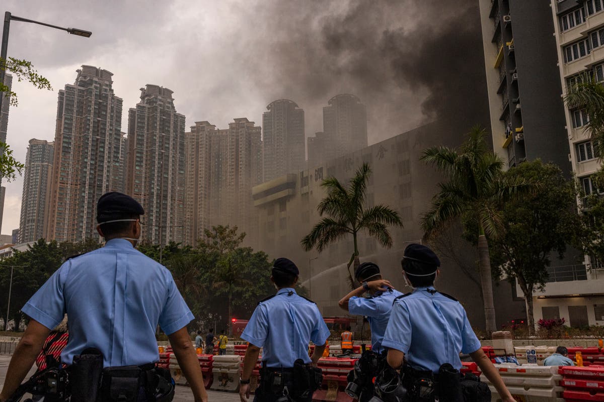 Four police officers wearing blue uniforms observe a fire engulfing a building in an urban area amidst dark smoke, with high-rise residential buildings and palm trees in the background.
