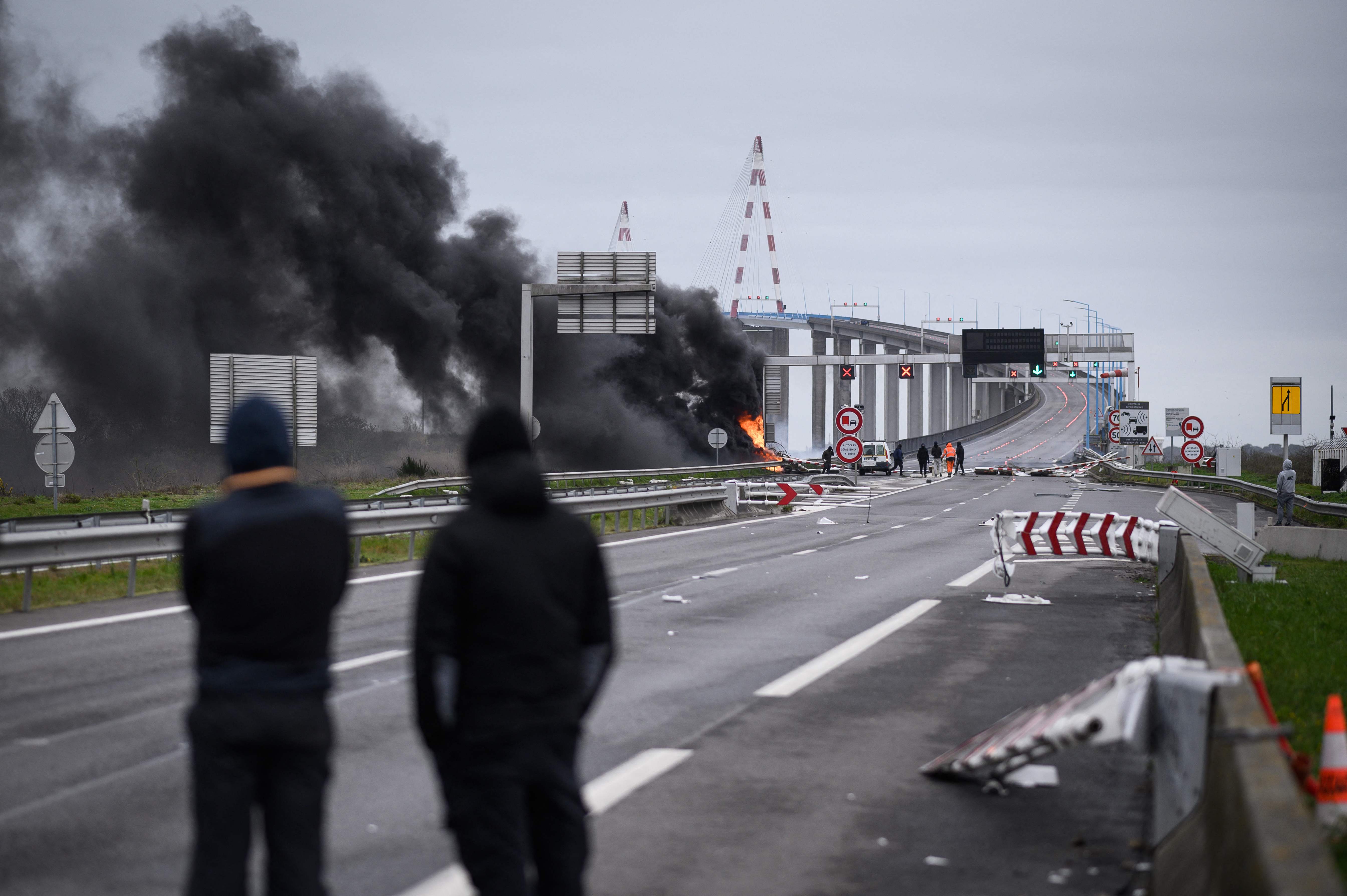 Protestors look on next to a damaged and burning road sign gantry at the entrance of the "Pont de Saint-Nazaire" (Saint-Nazaire bridge), the longest bridge in France