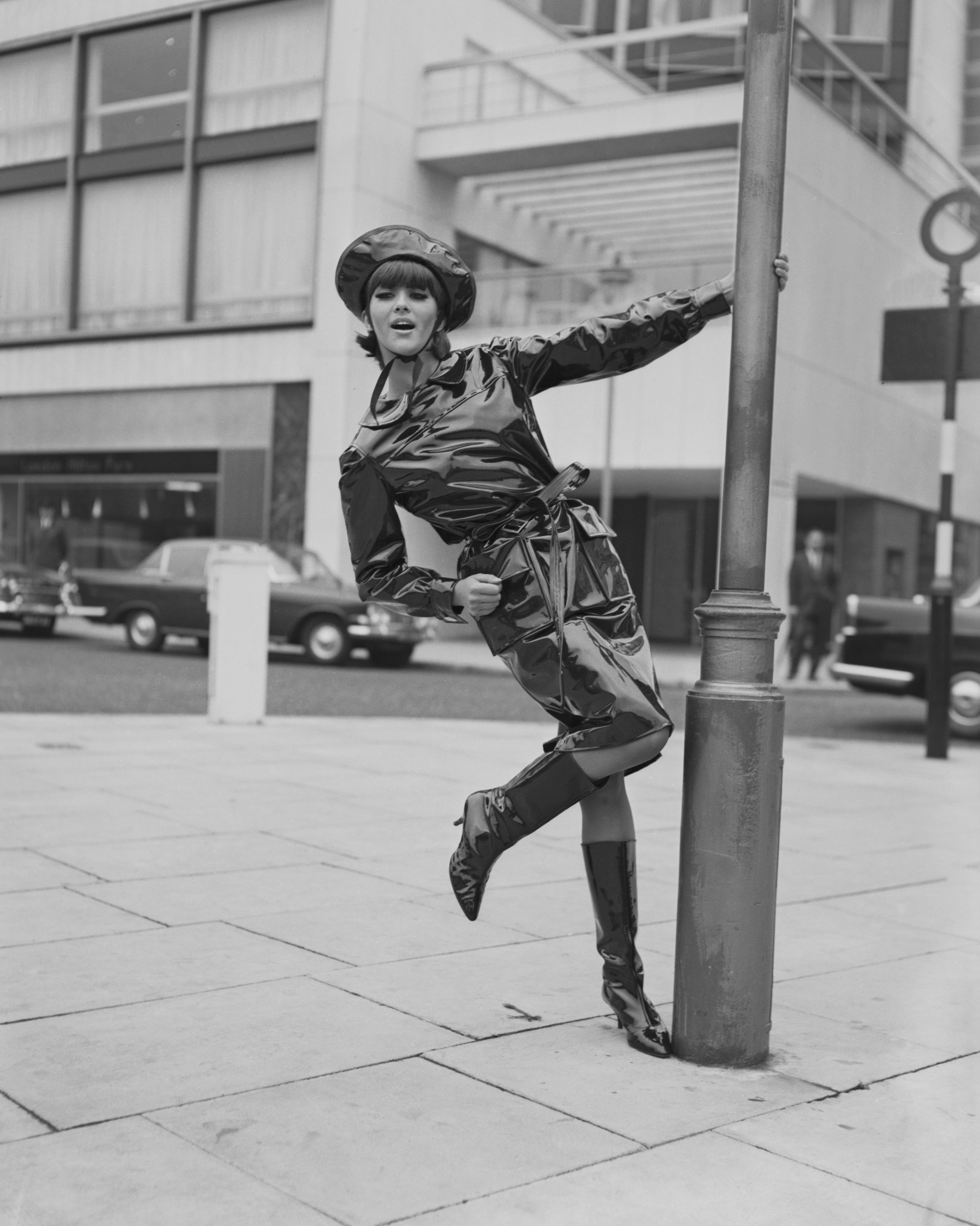 Model Jackie Bowyer swings on a lamppost wearing a black oilskin wet-weather outfit from Mary Quant, of matching coat and hat with shiny, high-heeled boots, London, 15th October 1963