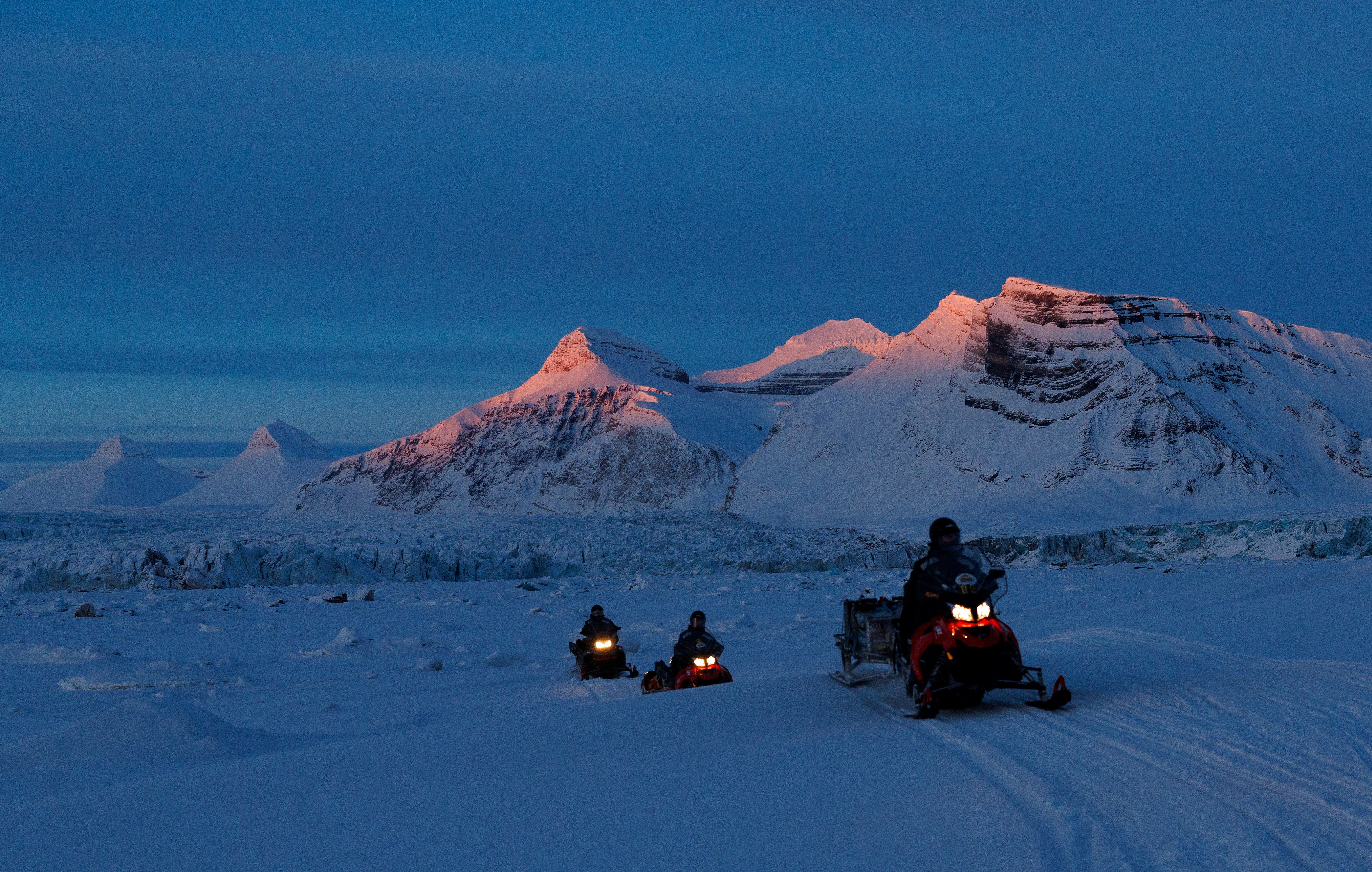 NPI (Norwegian Polar Institute) scientists ride their snowmobiles as the sun sets on Kongsfjord and Kronebreen glaciers