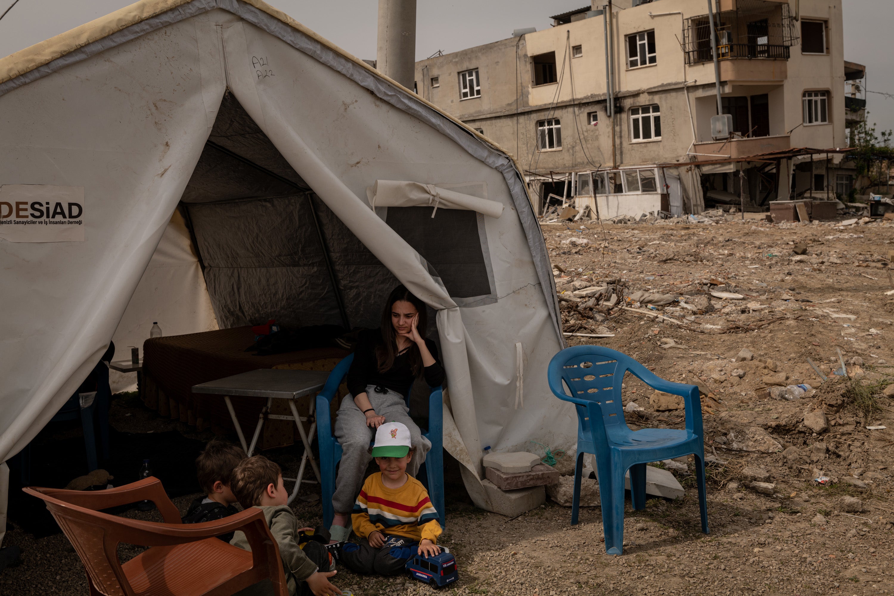 A young woman sits with children in front of their tent and near their former home