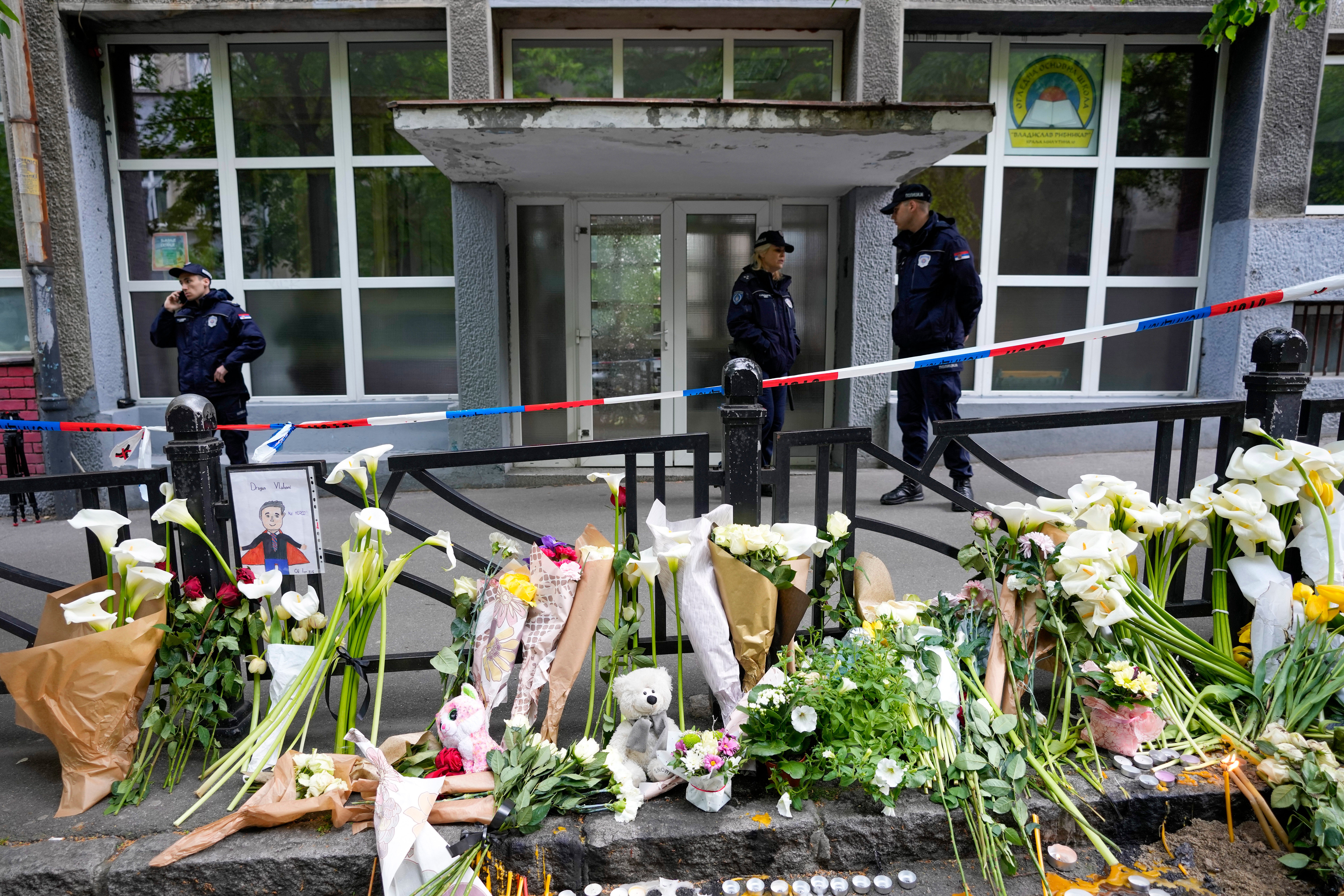 Police officers guard the Vladimir Ribnikar school in Belgrade – behind tributes to those killed
