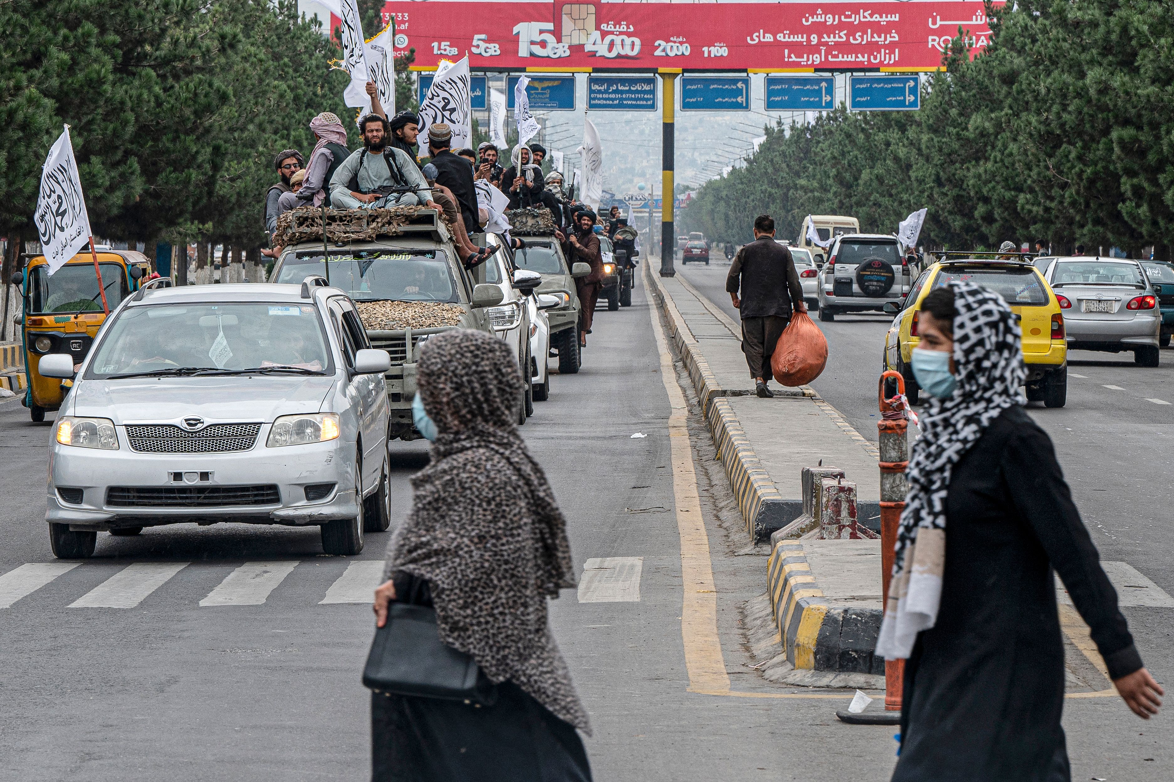 <p>Afghan women cross a road as Taliban fighters ride in a convoy to celebrate their victory day near the US embassy in Kabul</p>