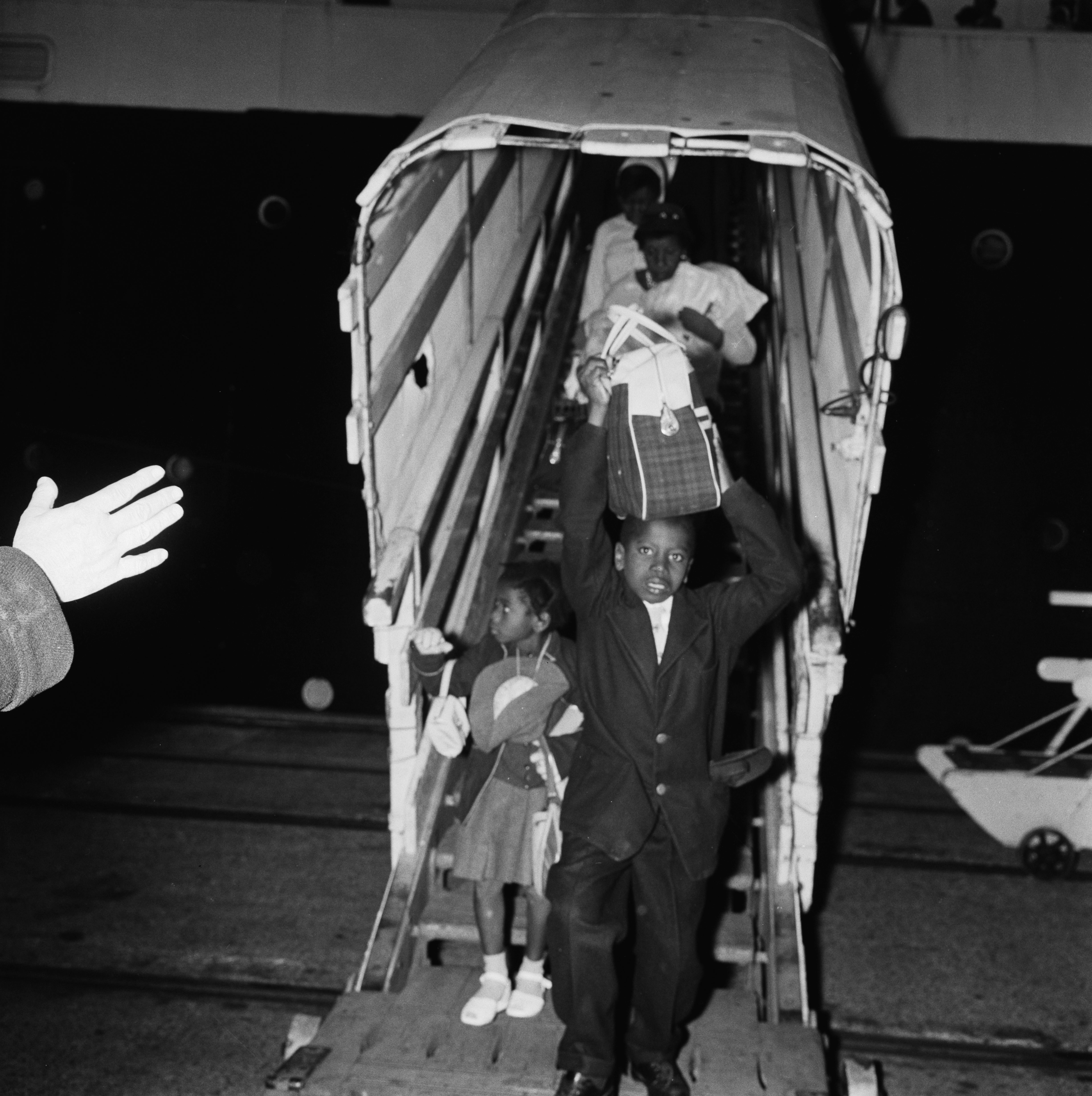 A boy holding a piece of luggage gets off a ship followed by a girl and two women at an unspecified port in 1961