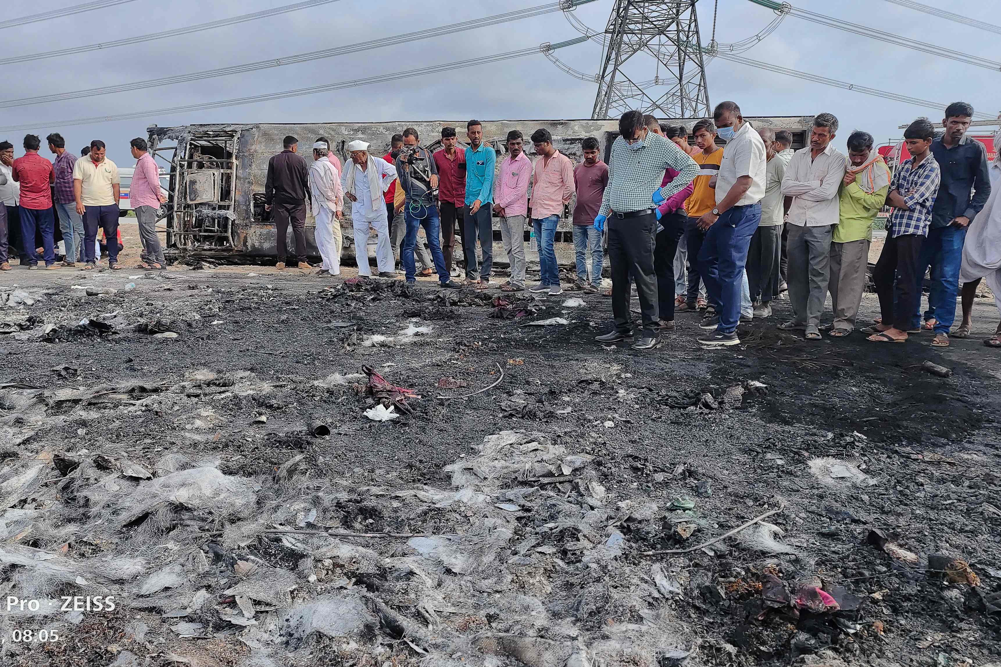 People gather next to the remains of a bus (background) that caught fire along the Samruddhi Expressway