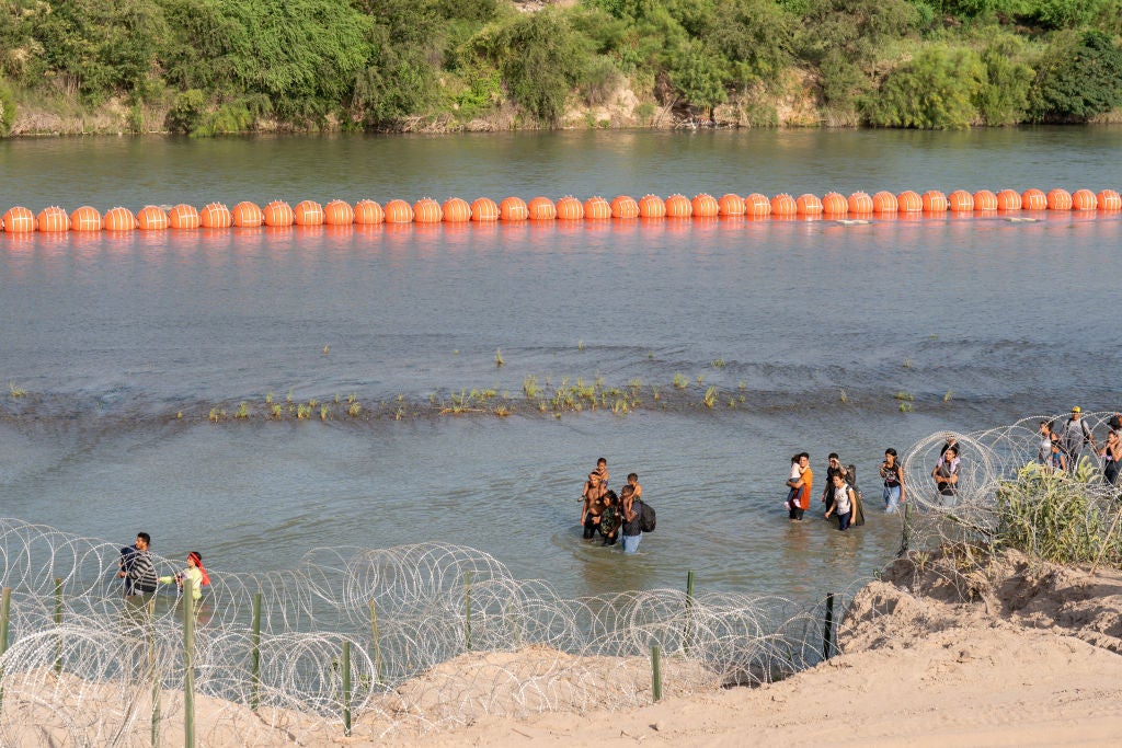 Migrants walk by a string of buoys placed on the water along the Rio Grande border with Mexico in Eagle Pass, Texas