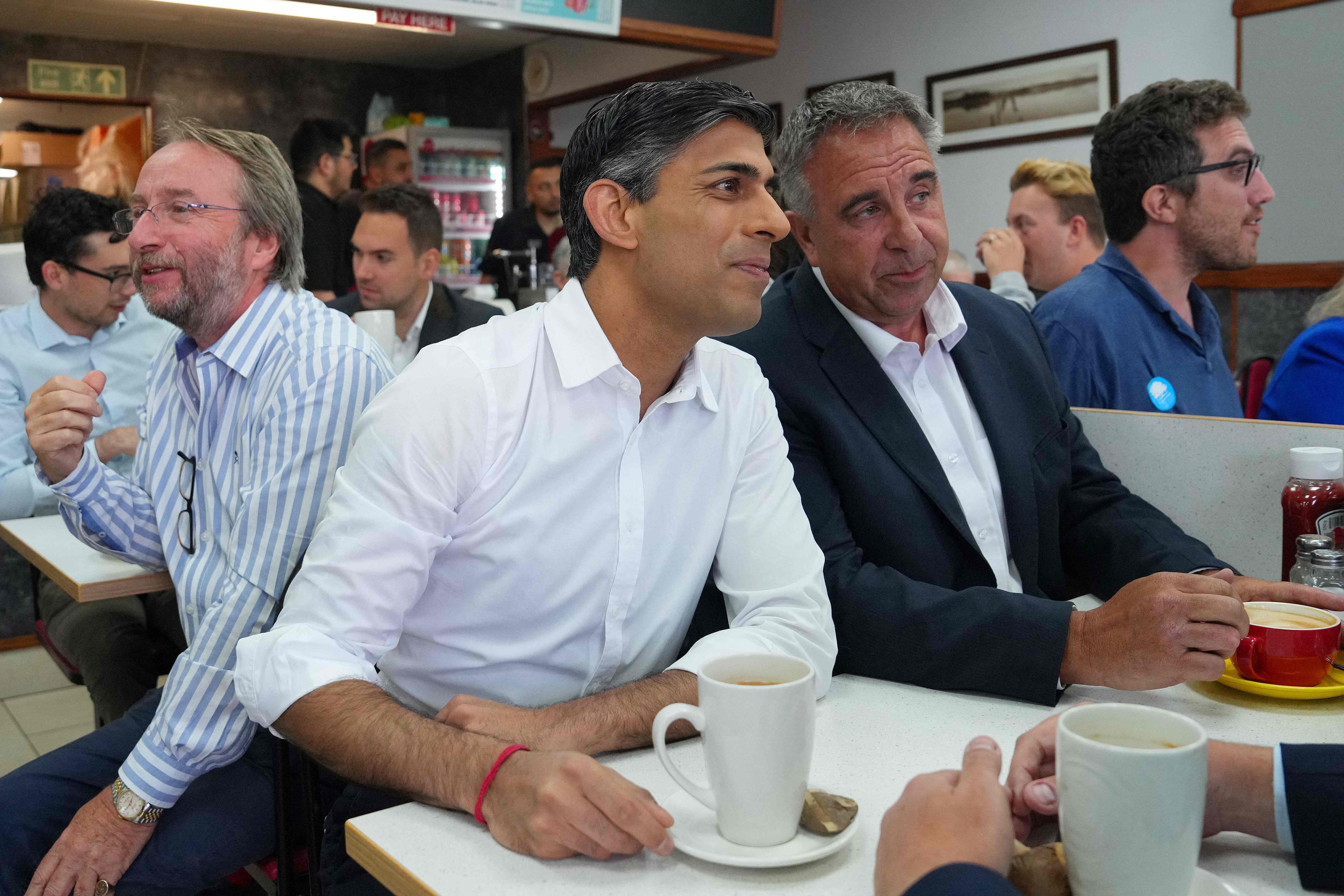 Britain's Prime Minister Rishi Sunak (C) sits with newly elected Conservative MP Steve Tuckwell (R) at a cafe in Ruislip