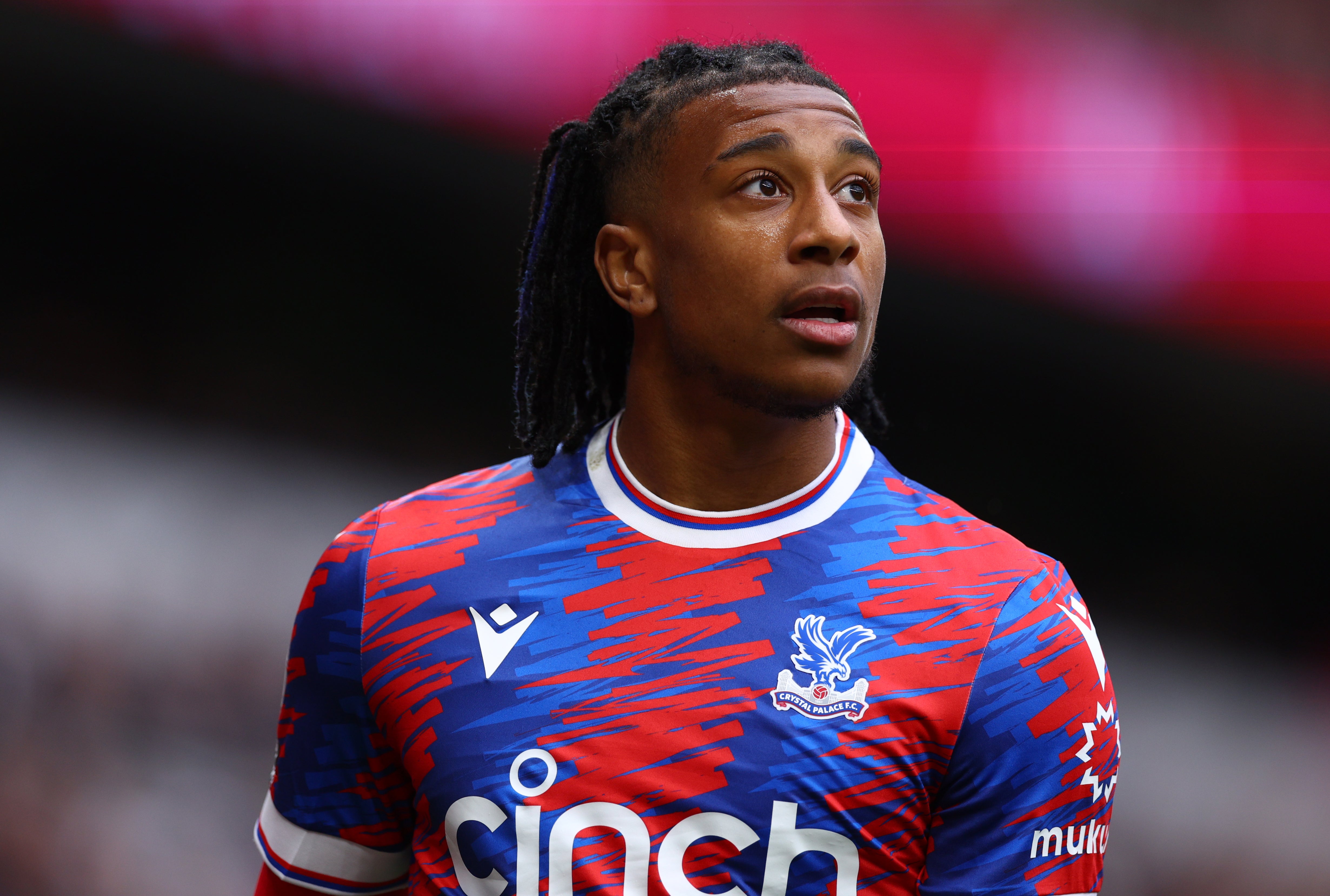  A young male football player with long dreadlocks looks up while playing a match on a field. The player is wearing a blue and red jersey with white shorts and socks.