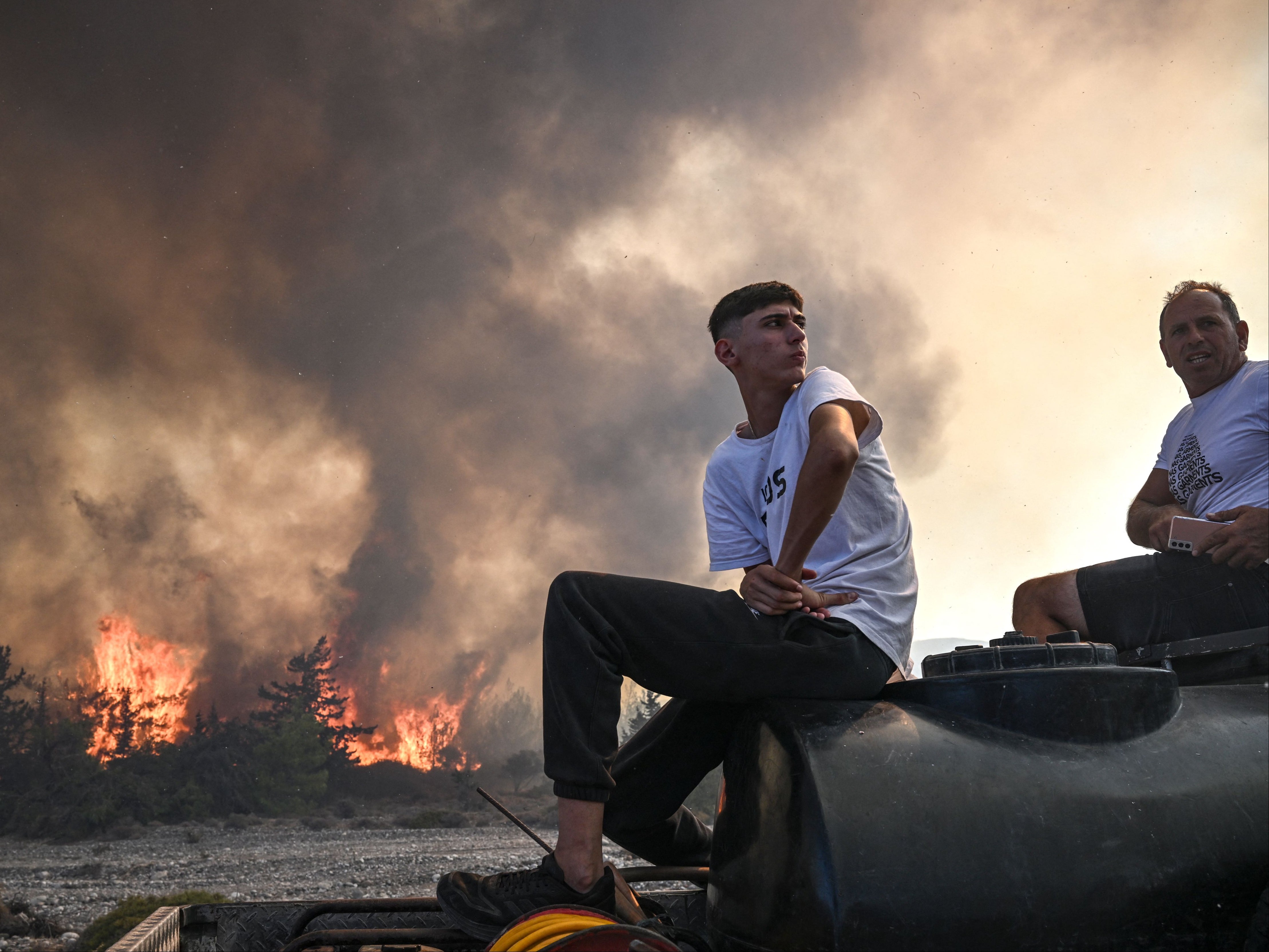 Men sit on the back of their pick-up truck as wildfires rage in the background close to the village of Vati on the Greek island of Rhodes on 25 July