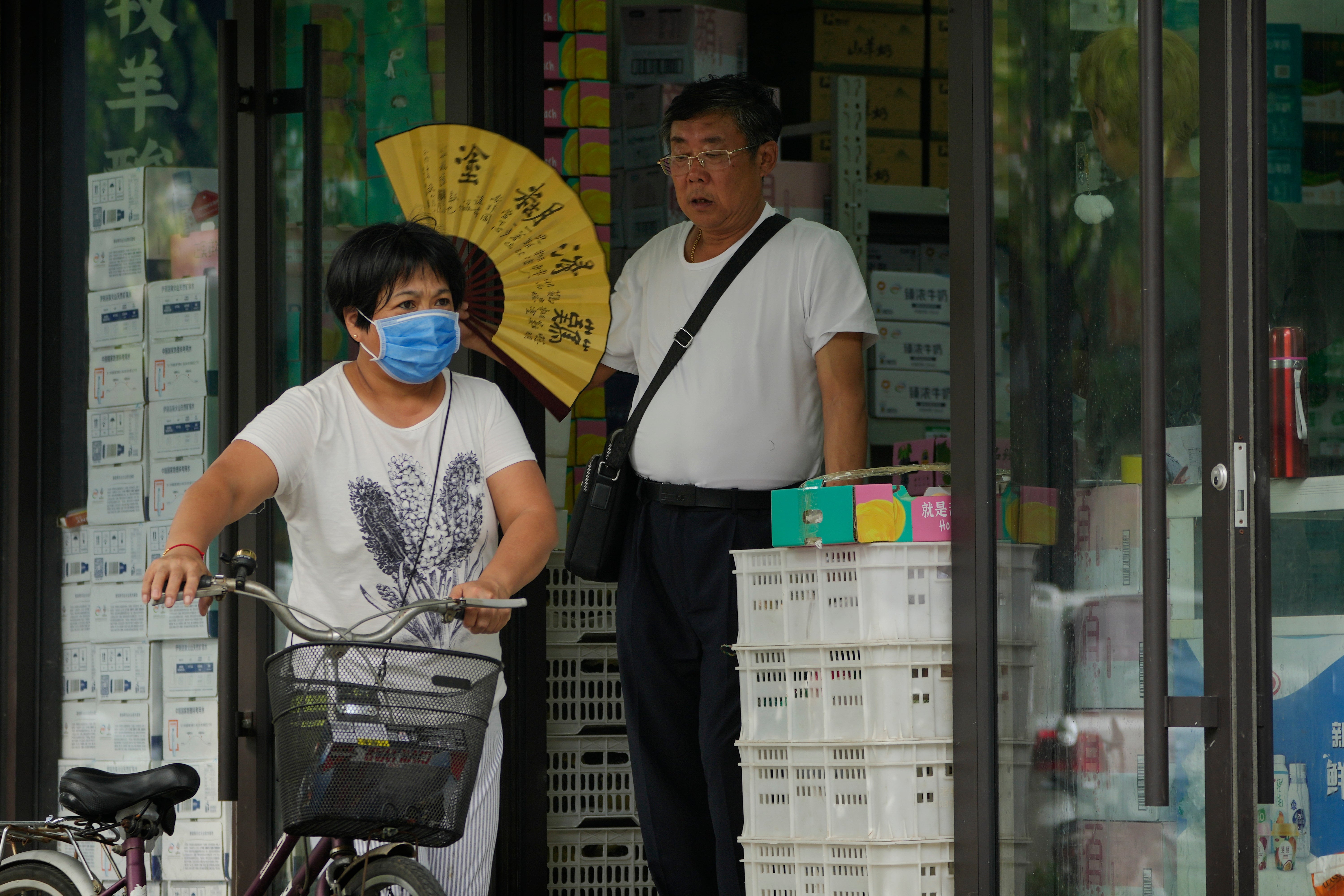 A woman pushes a bicycle past a man cooling himself with a fan at a store on a sweltering day in Beijing, July 24, 2023