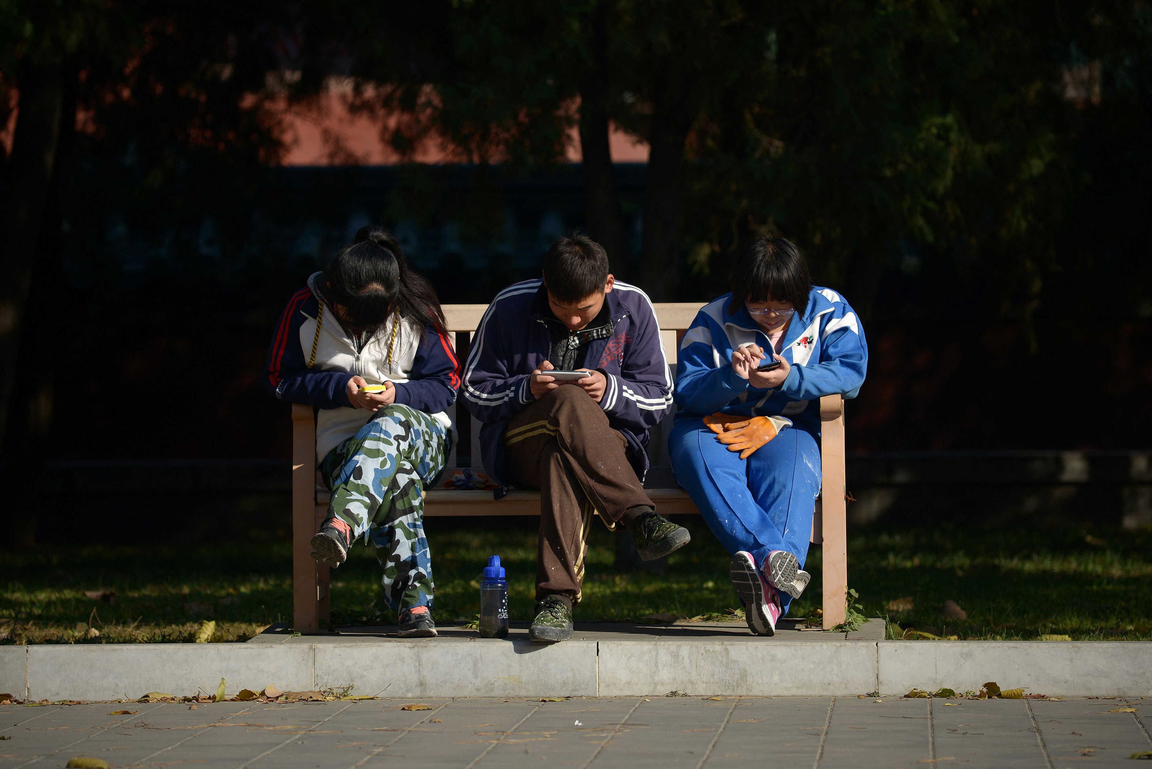 A group of young Chinese people play on their mobile phones as they rest on a bench at a park in Beijing