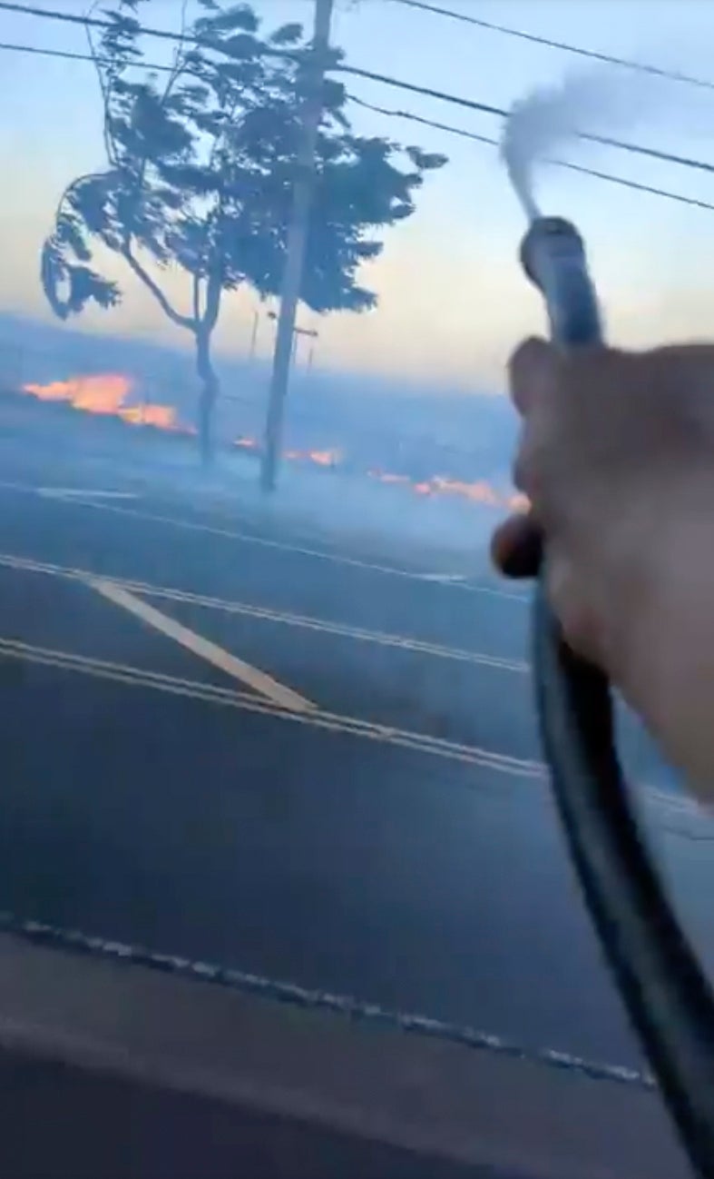 In this image from video posted by Shane Treu on 8 Aug 2023, he uses a garden hose to spray water during fires caused by snapped electrical cables on the Hawaiian island of Maui