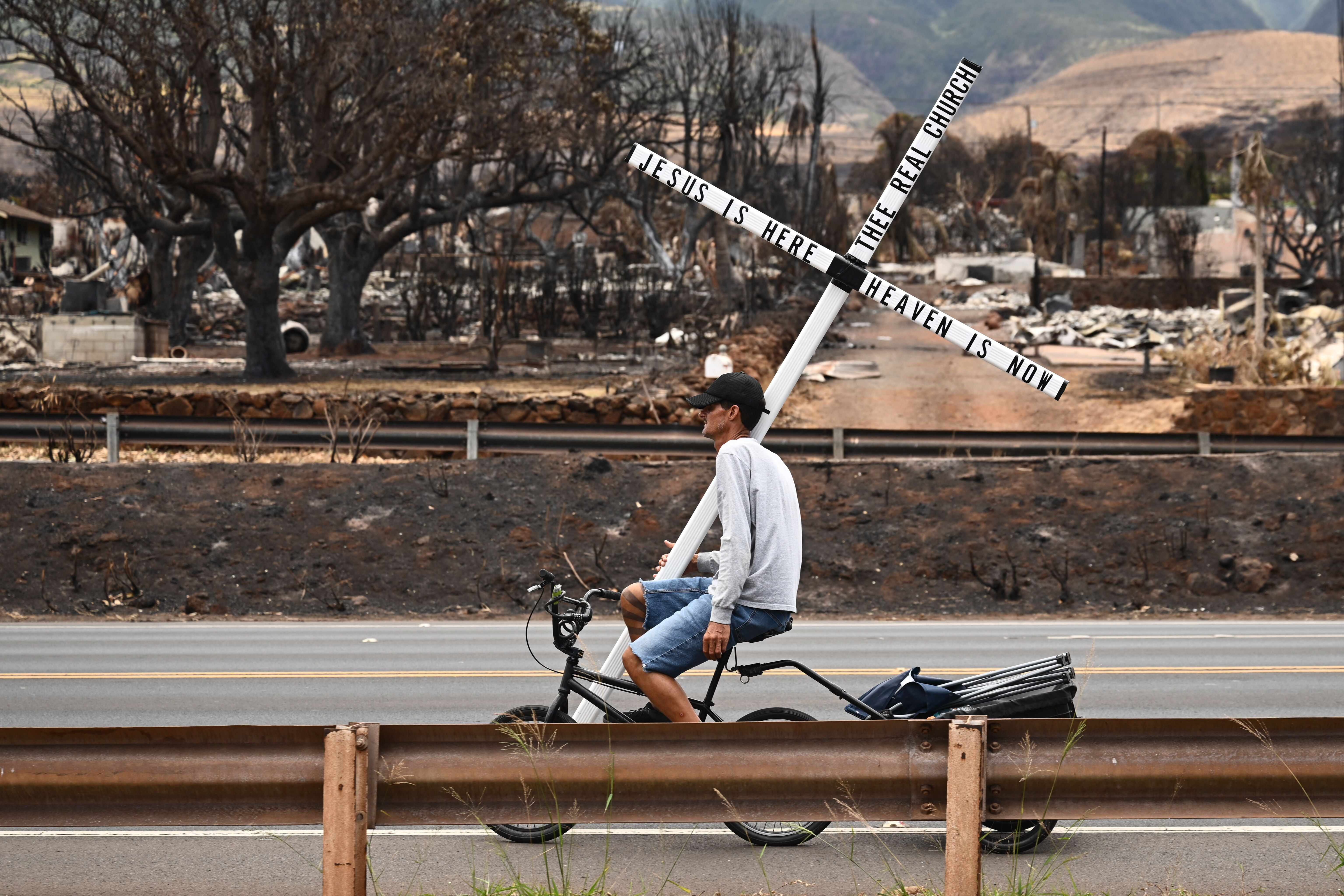 <p>A man on a bike carries a cross in the north side of Lahaina, Hawaii</p>