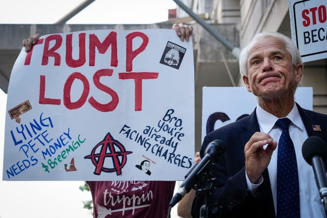 <p>A protester holds a sign behind former White House official Peter Navarro as he speaks outside of a federal courthouse in Washington DC</p>