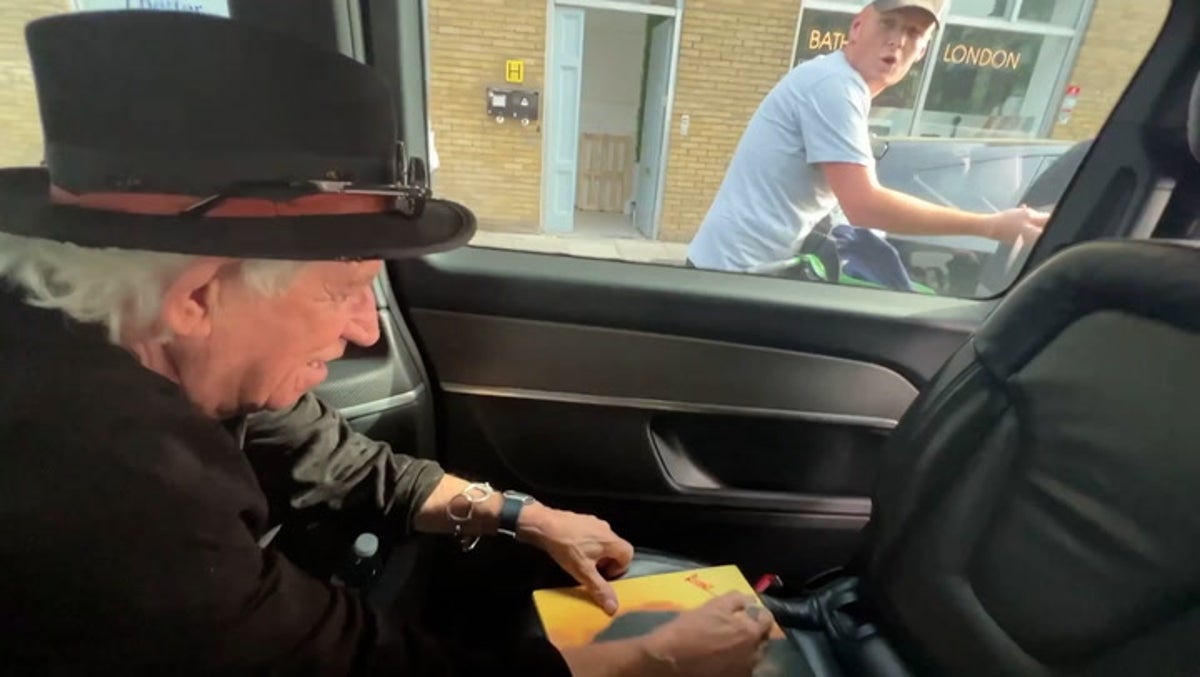 Rolling Stones’ Keith Richards jokes with cyclist as he signs autograph in back of taxi at traffic lights