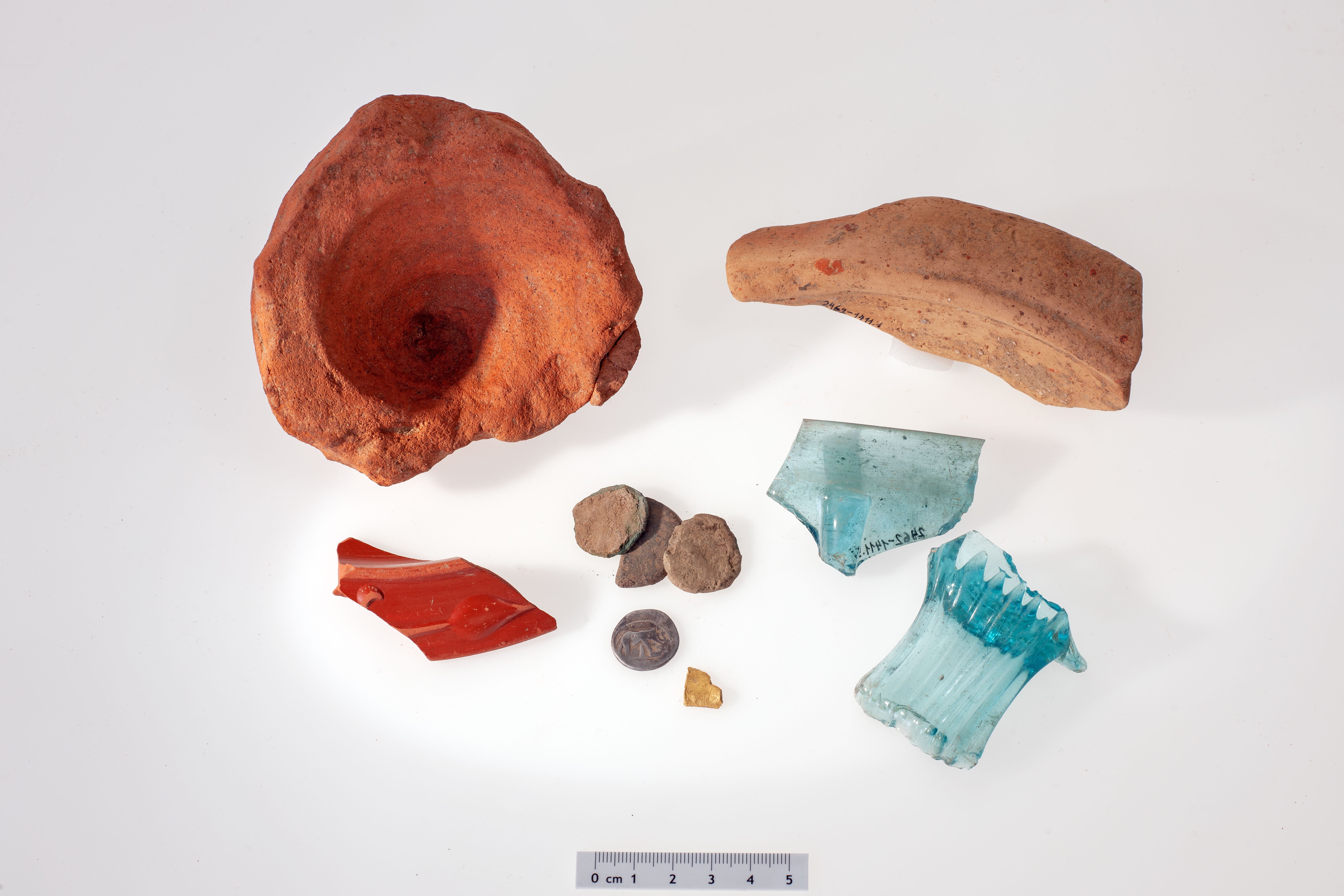 Small selection of Roman finds (from top left to bottom right): An amphora base, the shard of a rubbing bowl, the edge piece of a small bowl of Roman tableware with red coating (Terra Sigillata), four coins in found state, one of them made of silver by Julius Caesar, fragment of a gold object, pieces of a square bottle and a rib bowl made of blue glass