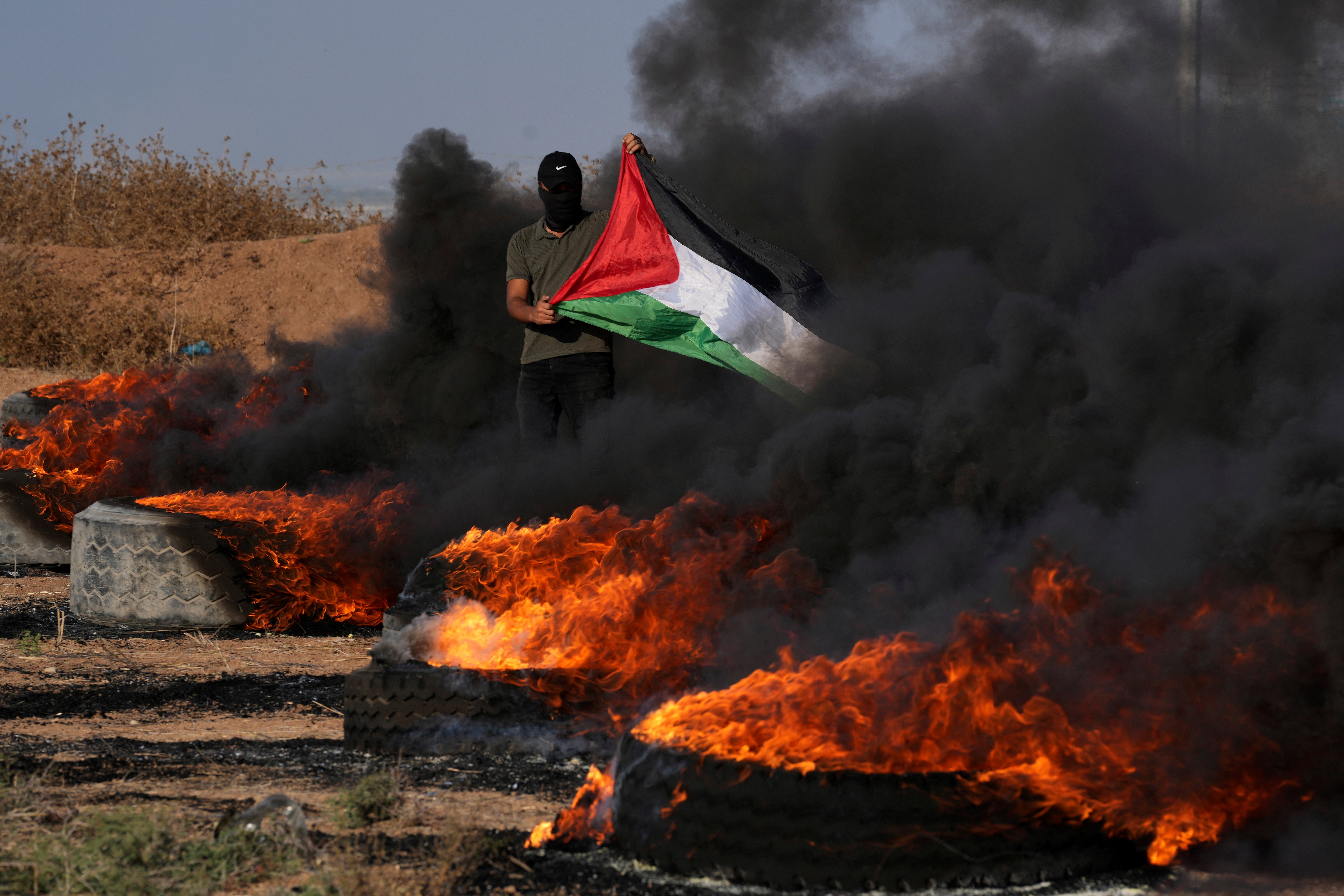 A masked Palestinian protester waves his national flag next to burning tires during clashes with Israeli security forces along the frontier with Israel