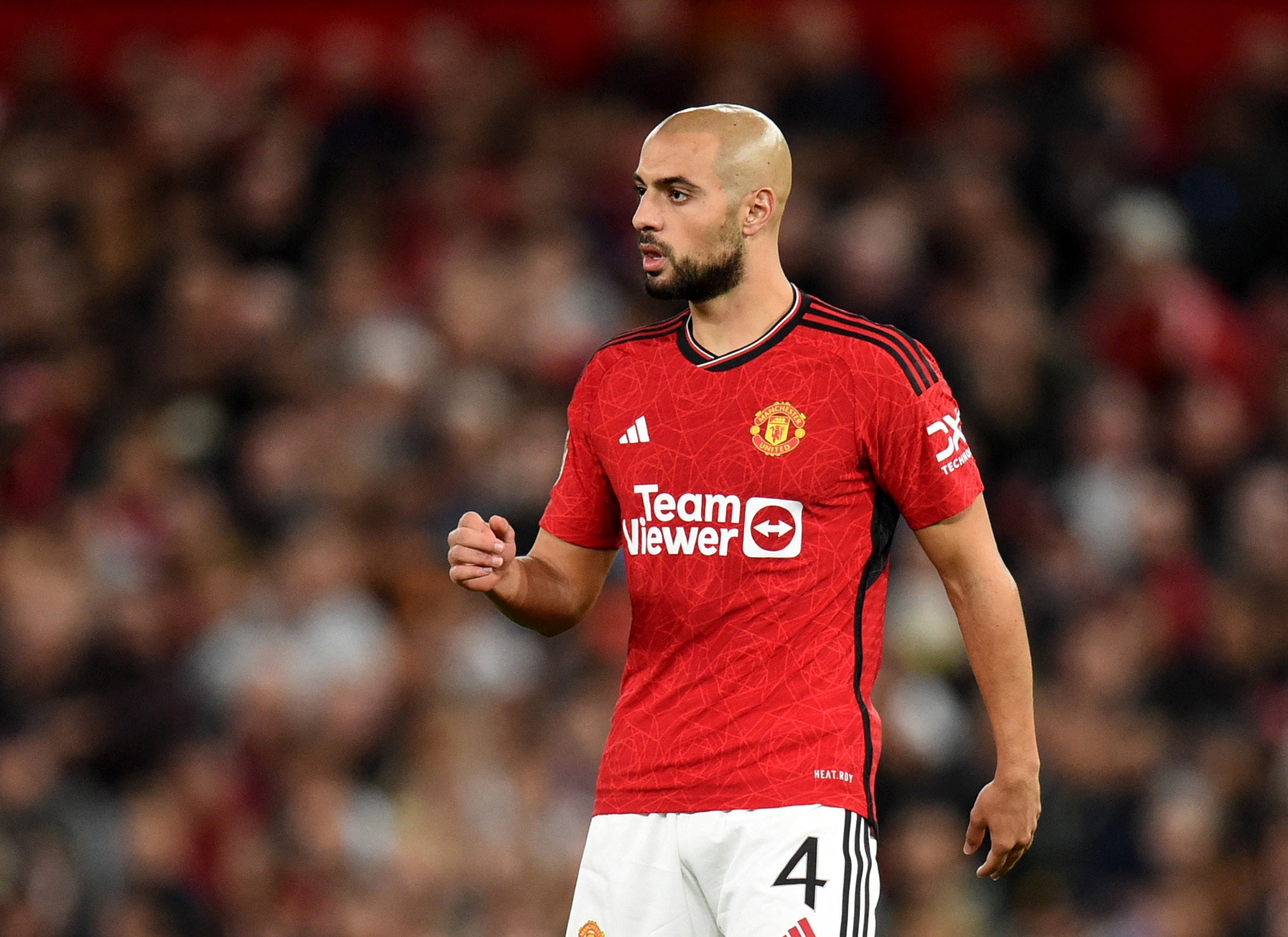 A bald Sofyan Amrabat is playing football for Manchester United, wearing a red jersey with a white collar and white shorts.
