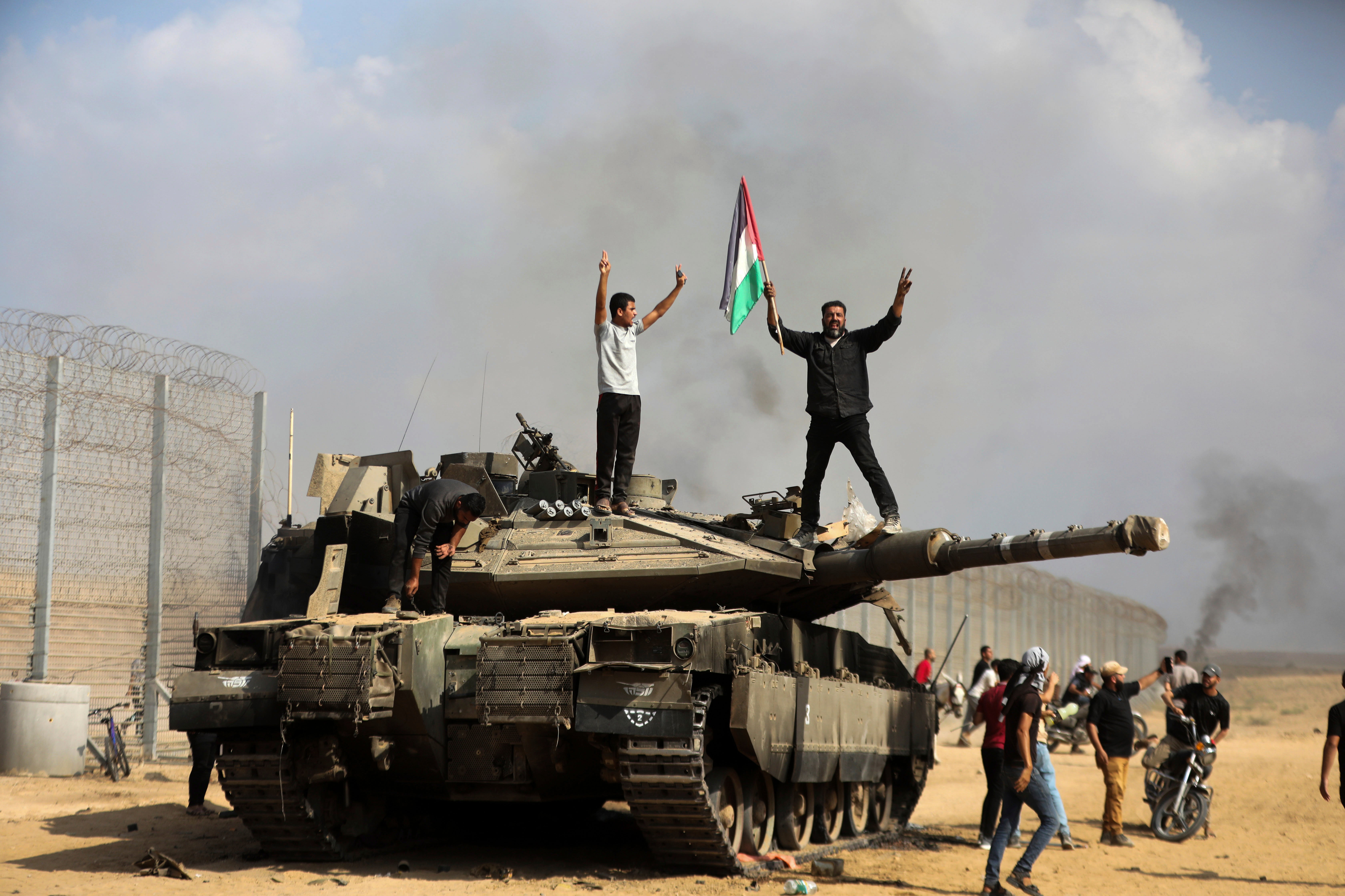Palestinians wave their national flag and celebrate by a destroyed Israeli tank at the Gaza Strip fence