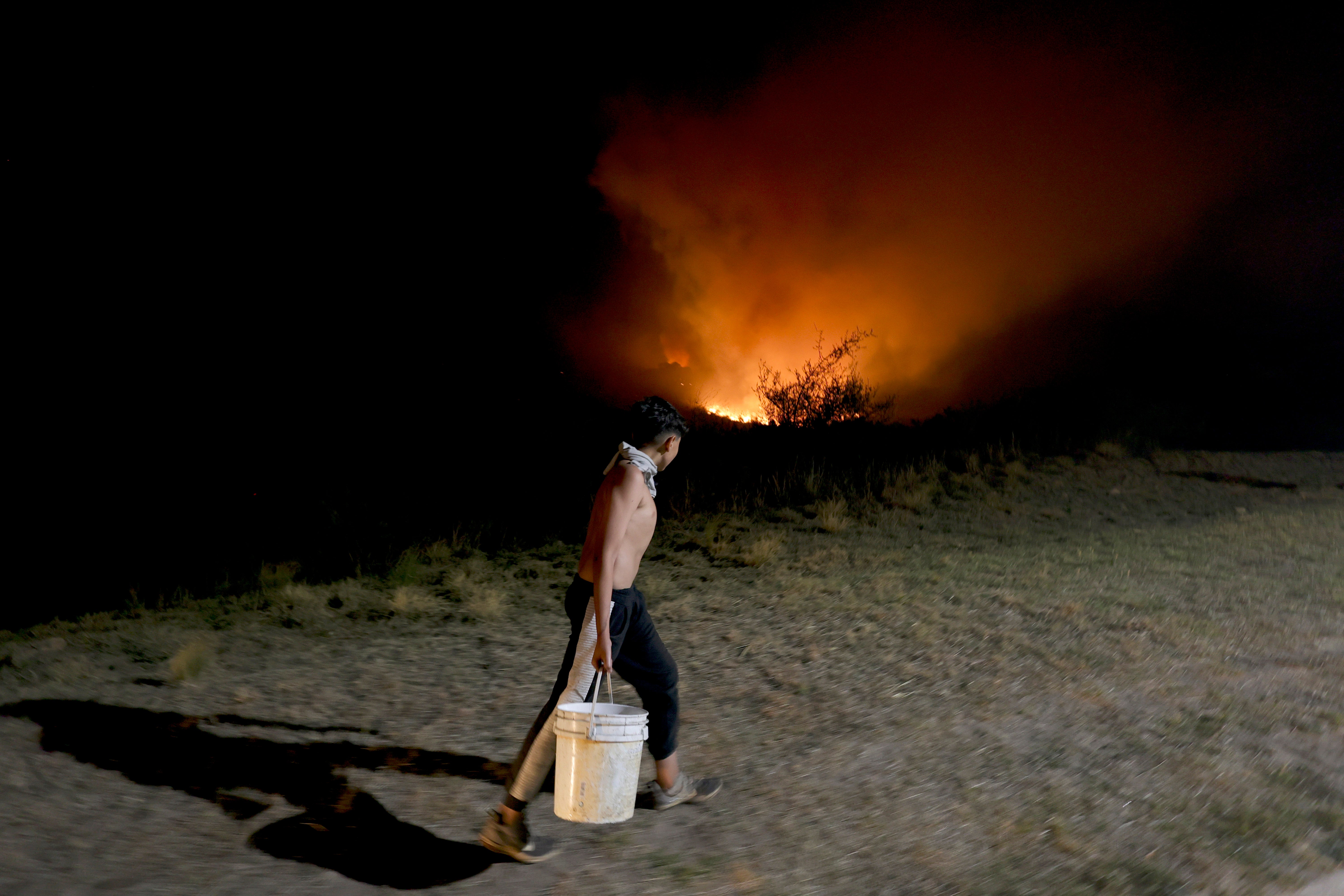 A man carries a bucket of water to help fight the forest fire behind him