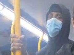 A masked man looks directly into a camera as he rides a west London bus