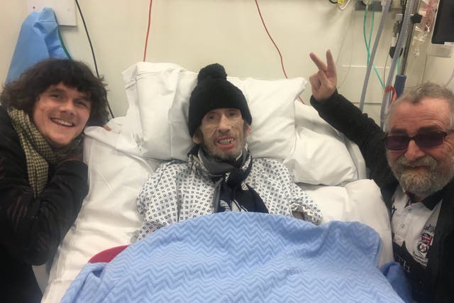 <p>Shane MacGowan smiling in a hospital bed wearing a scarf and bobble hat</p>