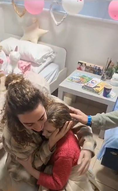 Emily, wearing a red knitted jumper, is embraced by a family member who strokes her face as the nine-year-old puts her arms around her