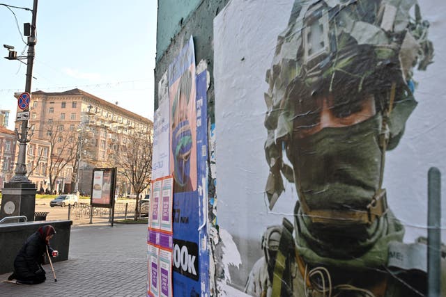 <p>An elderly woman kneels as she begs next to a poster of a Ukrainian soldier calling for donations to the army, in the center of the capital Kyiv</p>