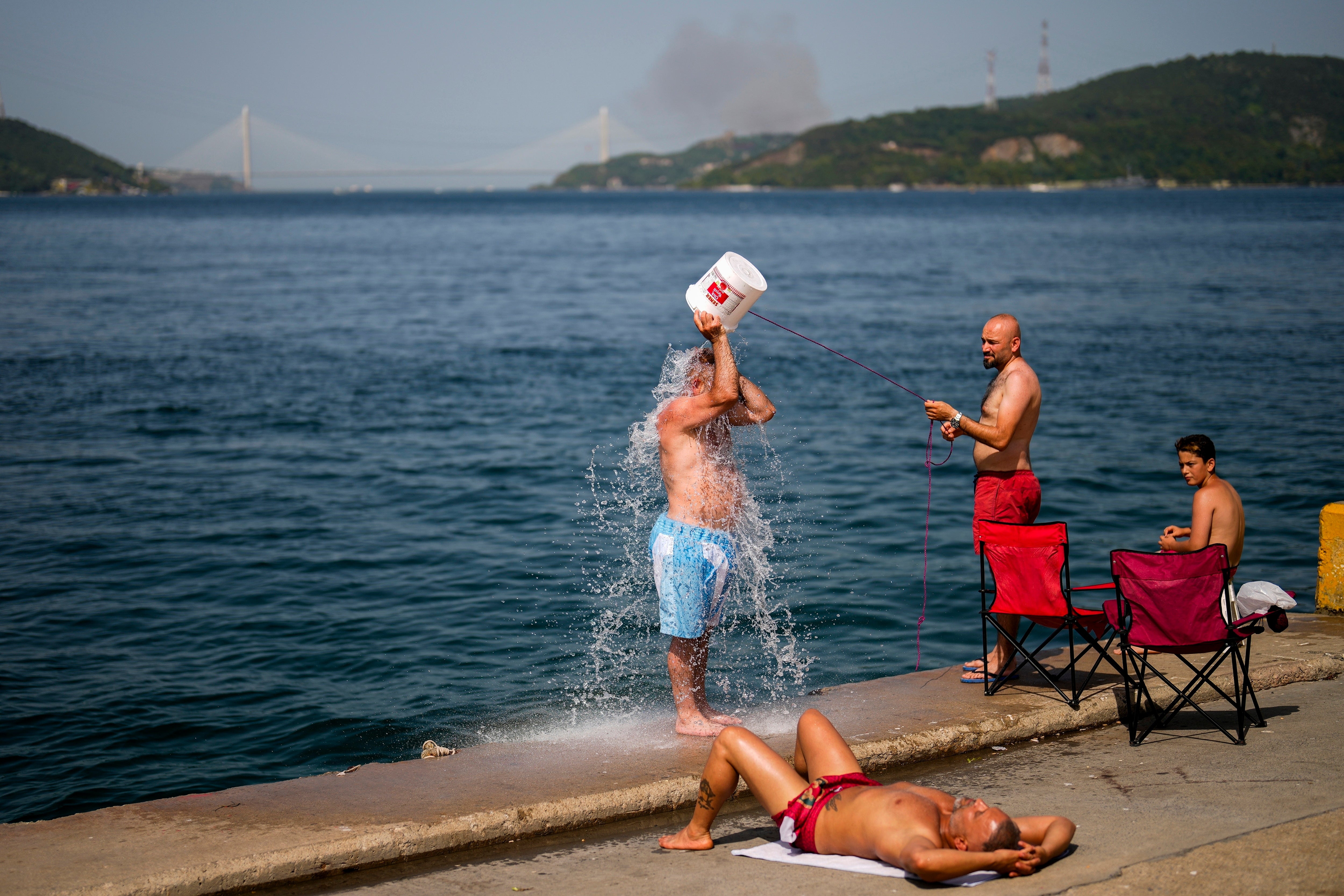 People cool off at the Bosphorus as a forest fire smoke rises, background, during a hot summer day in Istanbul