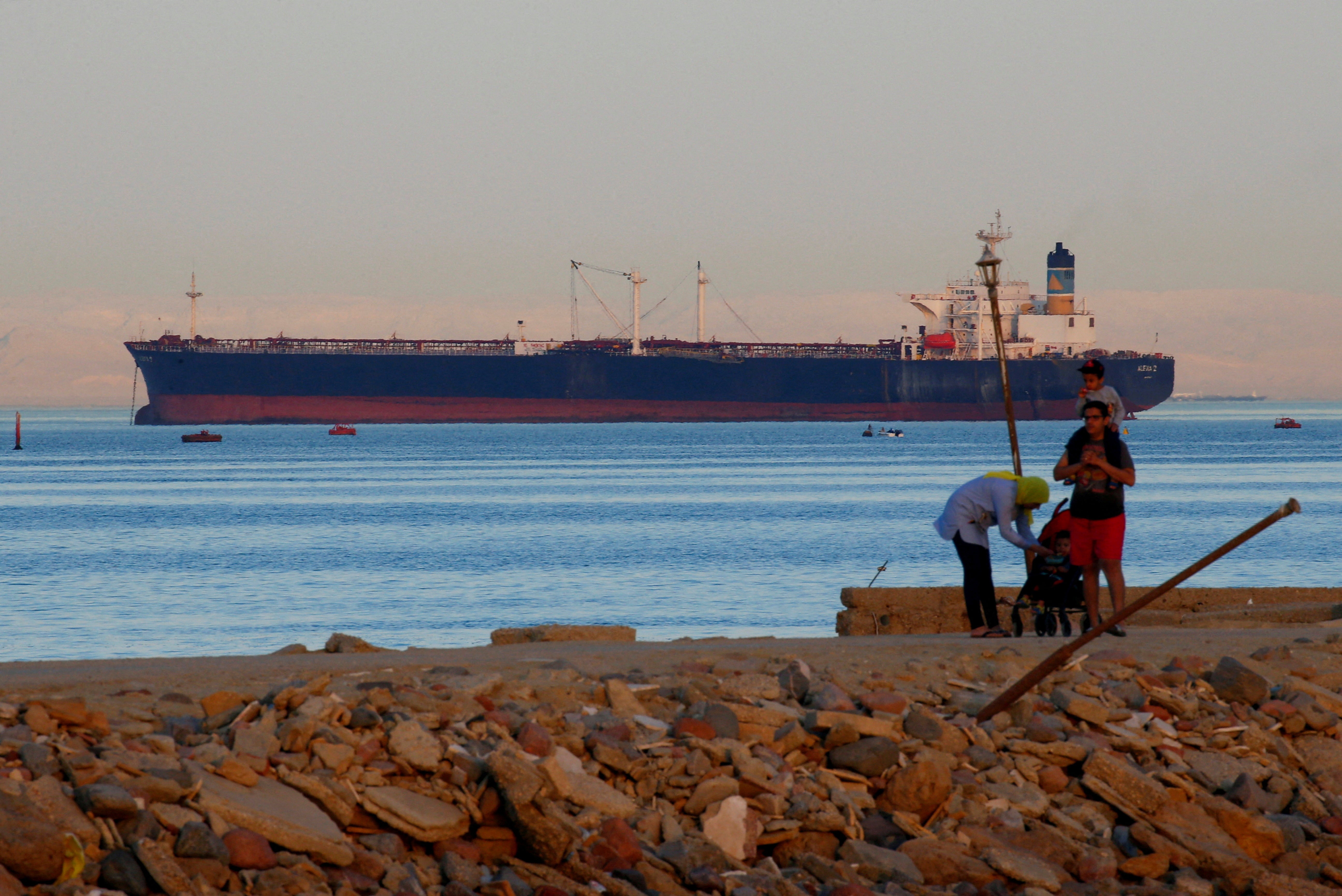 People walk on the beach as a container ship crosses the Gulf of Suez towards the Red Sea before entering the Suez Canal