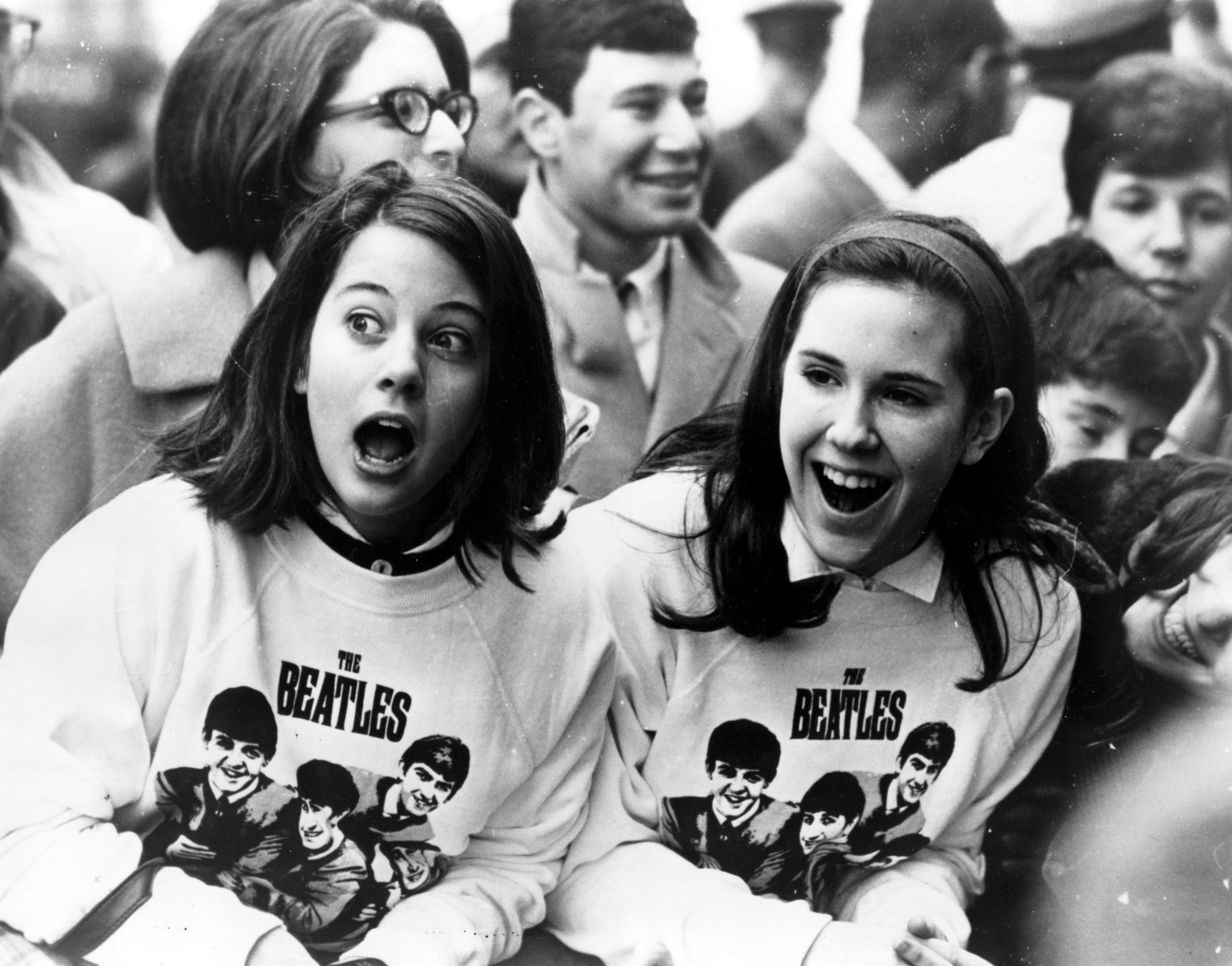 Two excited girls in Beatles sweatshirts among a crowd of fans in New York, welcoming the group as they arrive at the airport in February 1964