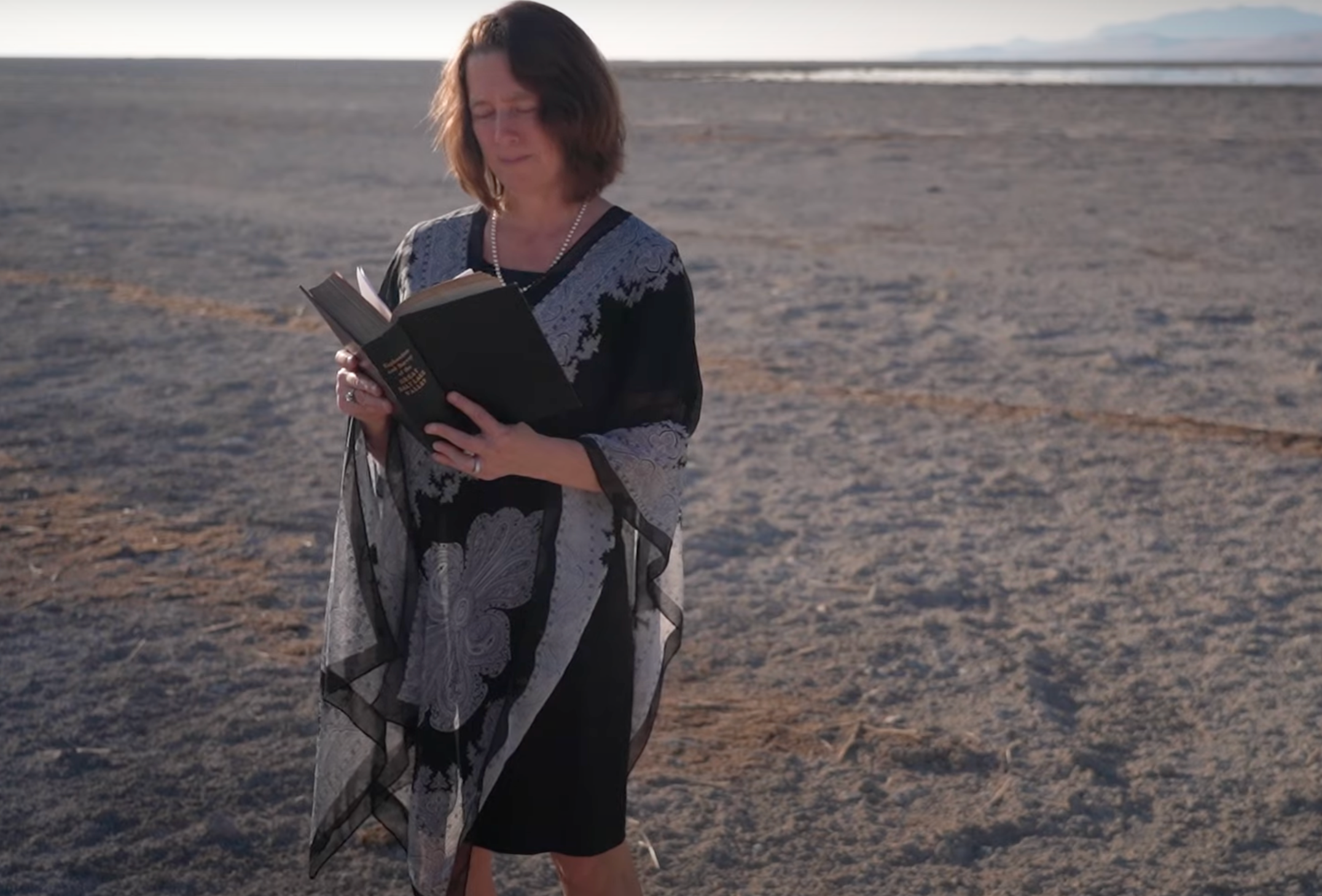 Bonnie Baxter reads an obituary standing on the dry ground where the Great Salt Lake has receded to raise awareness of its plight