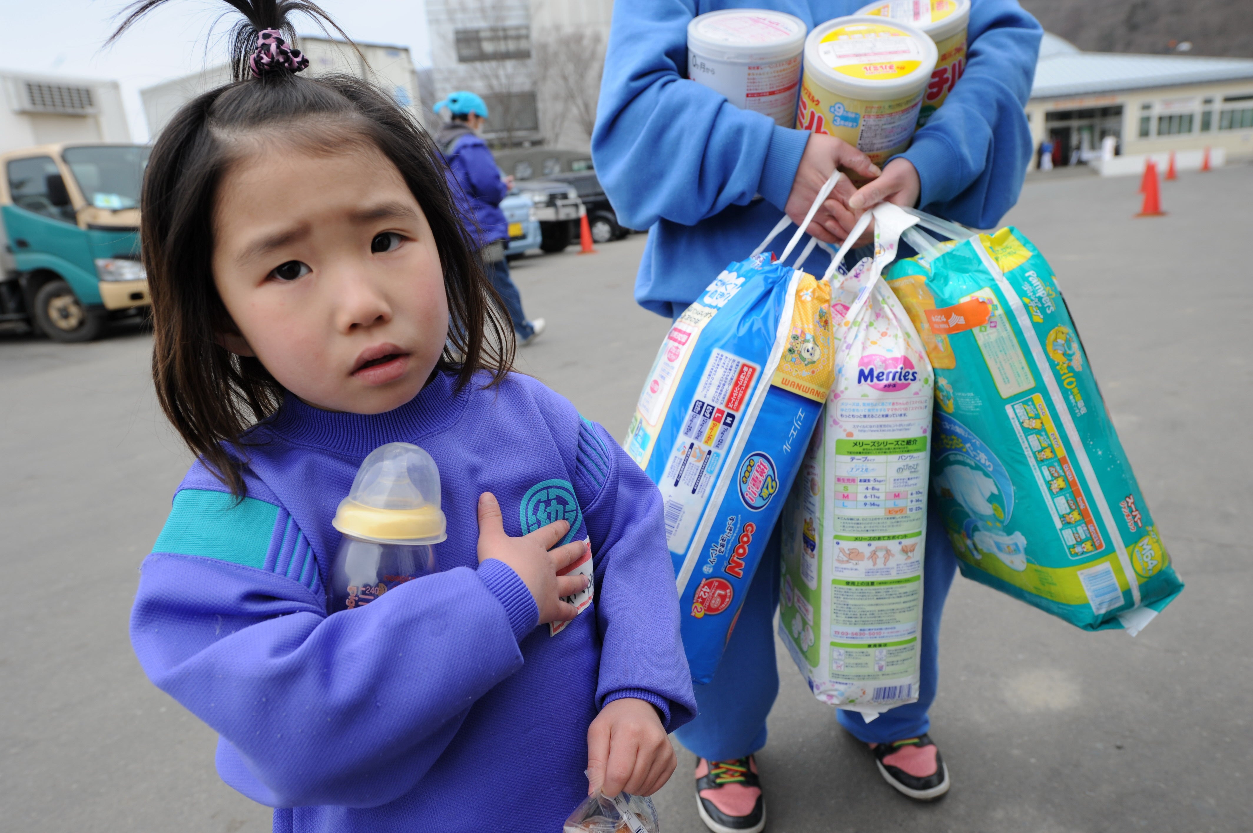 A little girl looks on while her mother carries baby diapers after collecting them from a distribution centre in the city of Kamaishi in Iwate prefecture