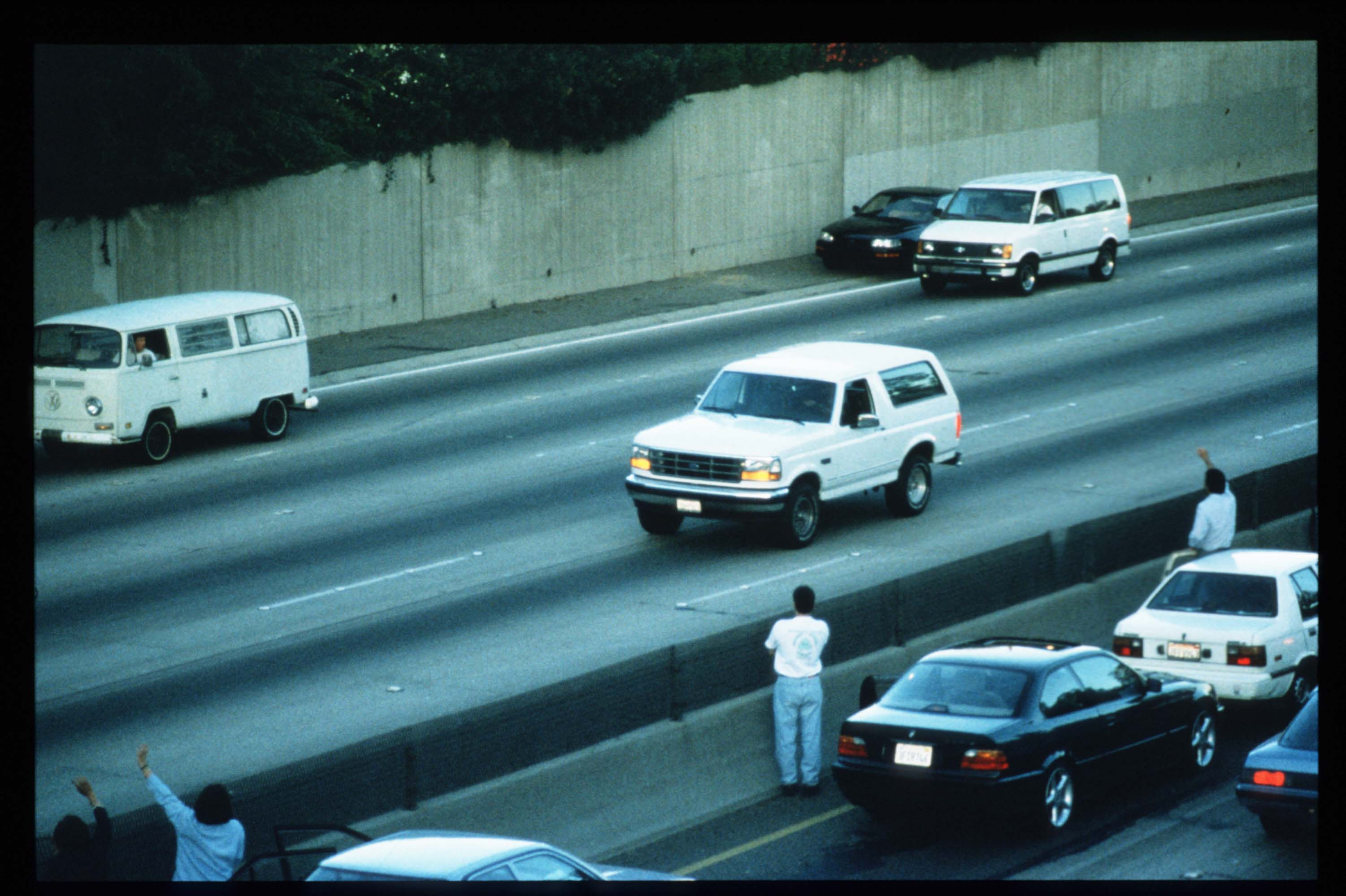 Motorists wave as police cars pursue the Ford Bronco (white, R) driven by Al Cowlings, carrying fugitive murder suspect O.J. Simpson, on a 90-minute slow-speed car chase June 17, 1994
