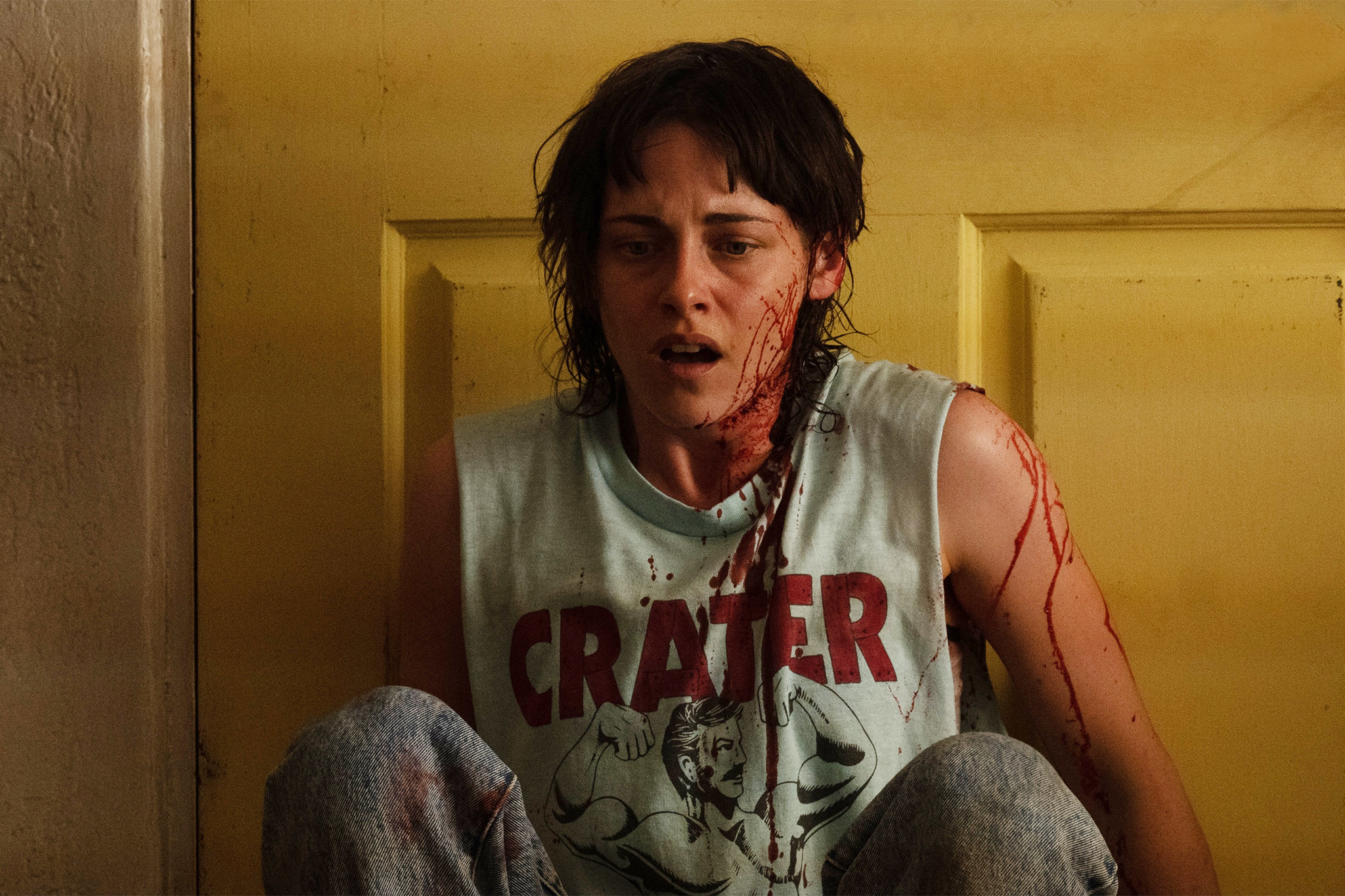 Sentada no chão e encostada na porta amarela, temos a personagem de Kristen Steart. Ela está em choque, coberta de sangue que não é dela. | Cena do filme "O Amor Sangra"