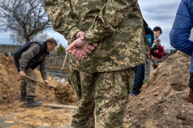<p>A Ukrainian soldier watches as grave diggers shovel soil into an open grave in Bucha, Ukraine</p>