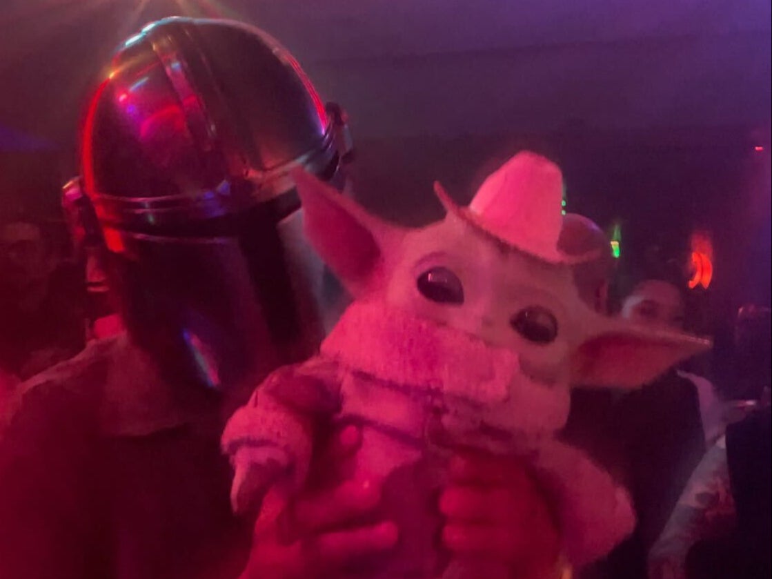 A man in a Mandalorian mask lifts up his baby Grogu doll that’s wearing a tiny sombrero atop his head.