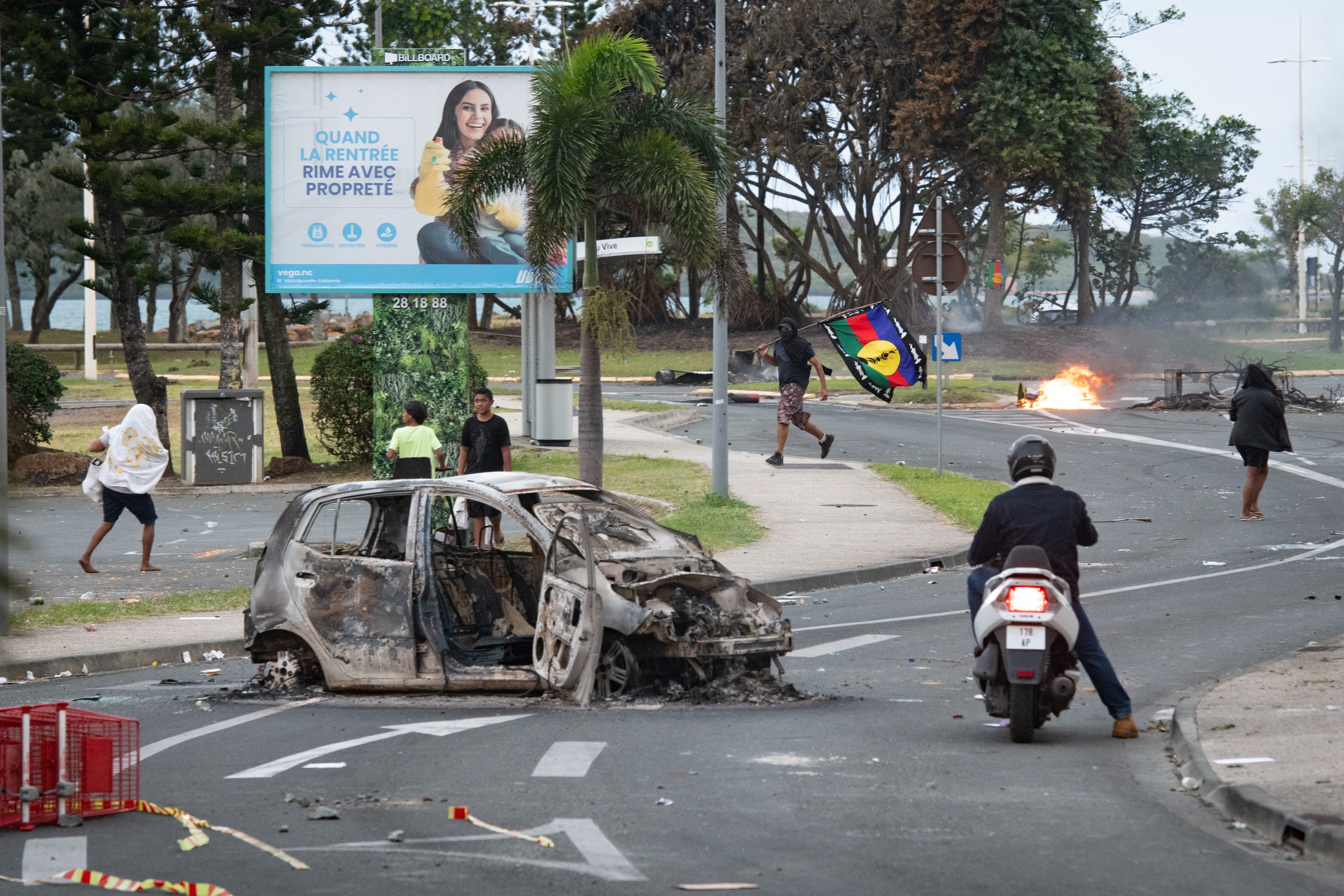 People walk next to a burnt-out car while a man (C) holds a flag of the Socialist Kanak National Liberation Front (FLNKS) after a supermarket was looted and shops vandalised in the N'Gea district of Noumea, on May 14, 2024