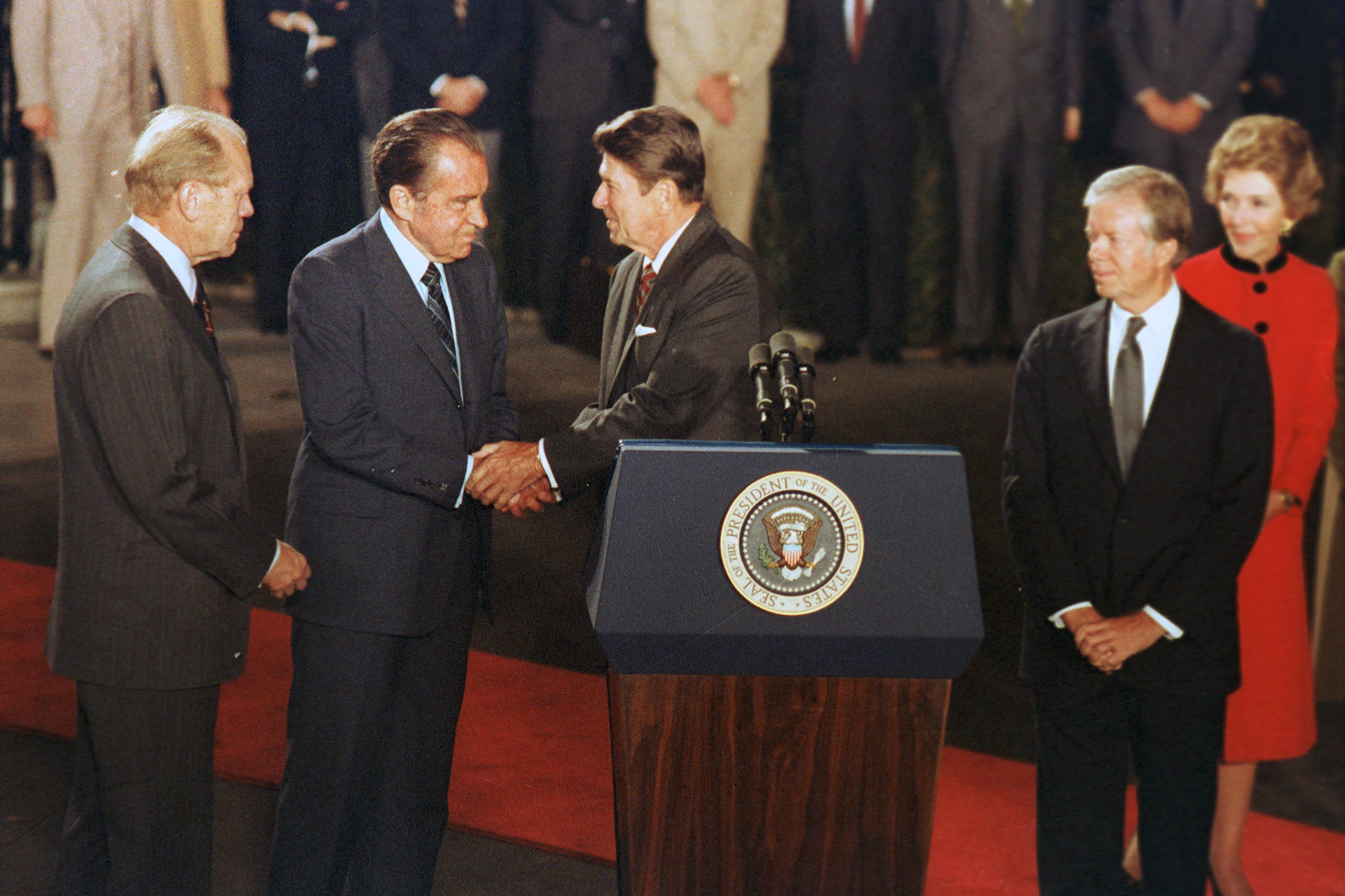 Presiden AS Ronald Reagan, di belakang podium, berjabat tangan dengan mantan Presiden Richard Nixon, sementara mantan Presiden Gerald Ford, kiri, dan Jimmy Carter, kanan, melihat ke Gedung Putih di Washington DC pada tahun 1981