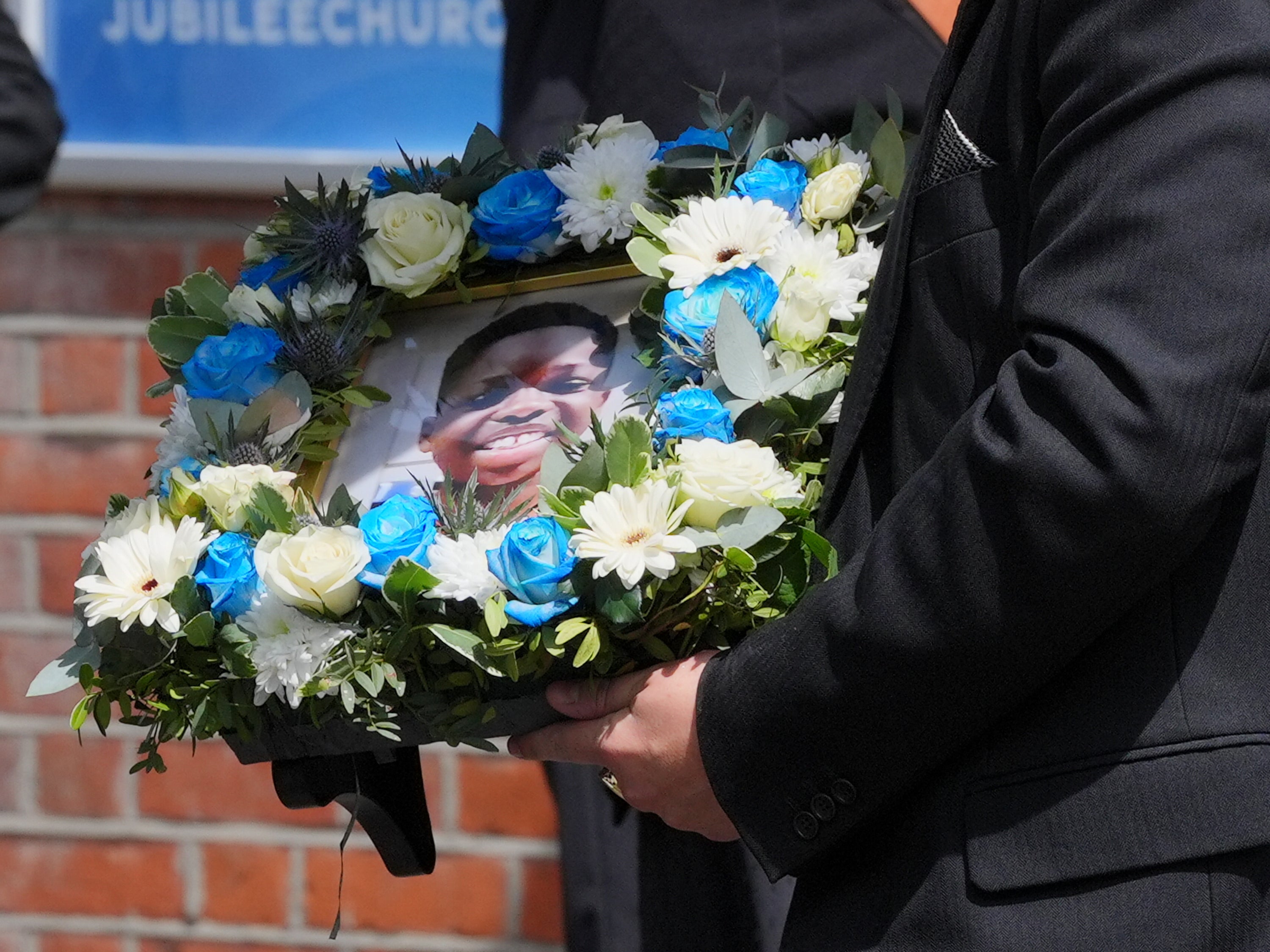 A funeral director carries a floral tribute inset with a photograph of Daniel Anjorin