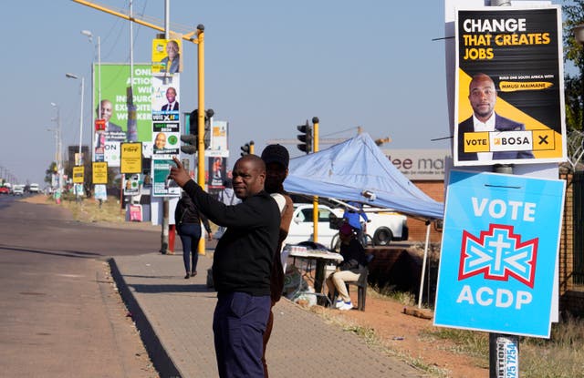 <p>A man waits for a taxi along a street lined with election posters in the Hammanskraal township, Pretoria</p>
