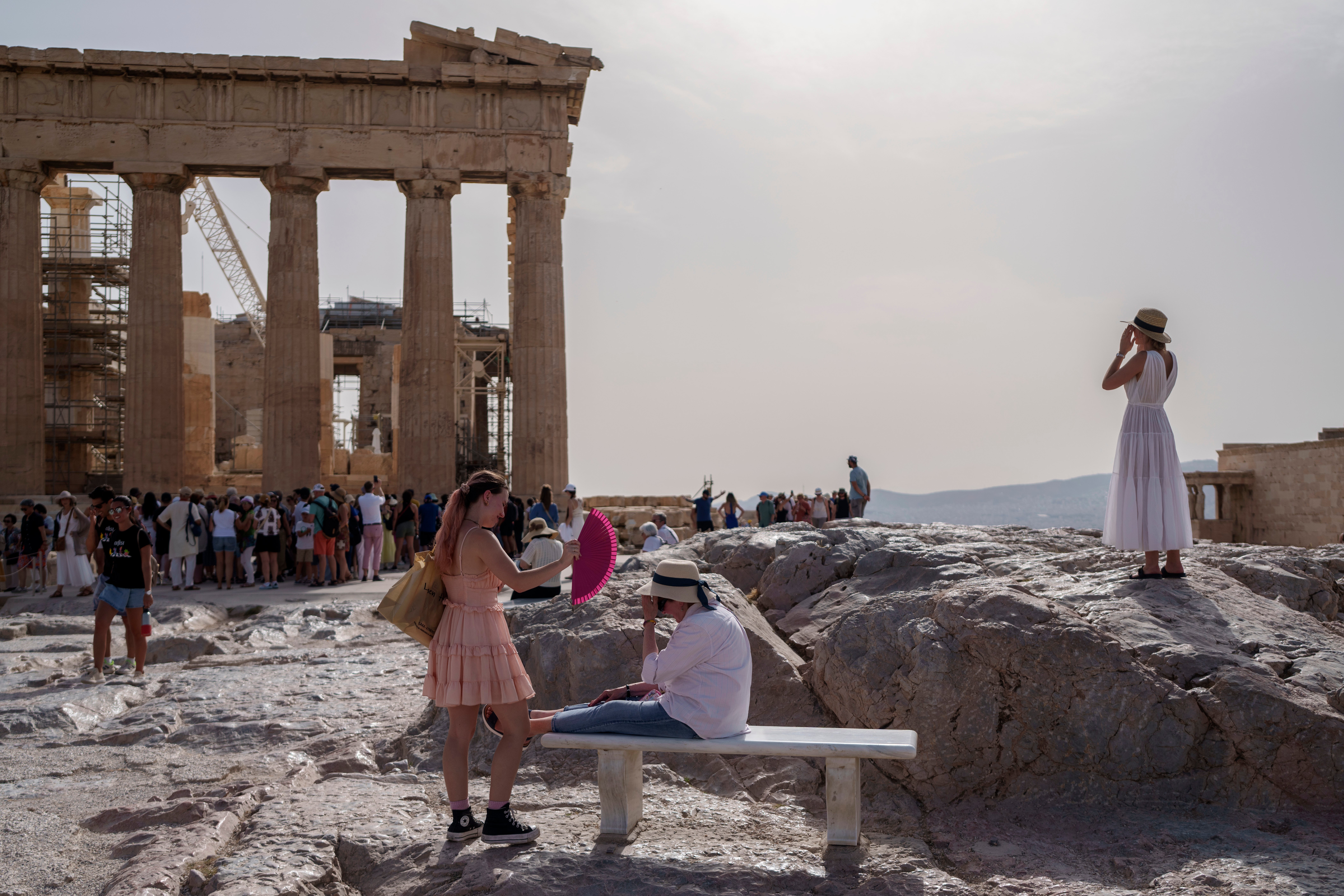 A tourist uses a hand fan to cool down another one sitting on a bench in front of the Parthenon at the ancient Acropolis, in Athens