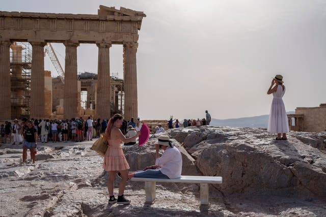 <p>A tourist uses a hand fan to cool down another one sitting on a bench in front of the Parthenon at the ancient Acropolis, in Athens</p>